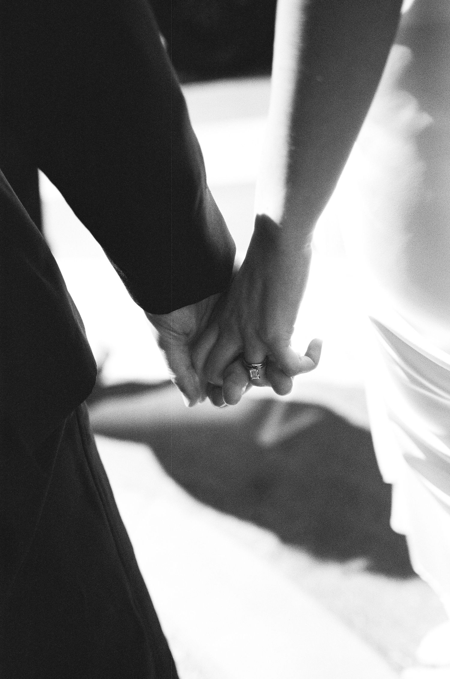 Black and white photo of a bride and groom holding hands, showcasing a diamond engagement ring on their wedding day.
