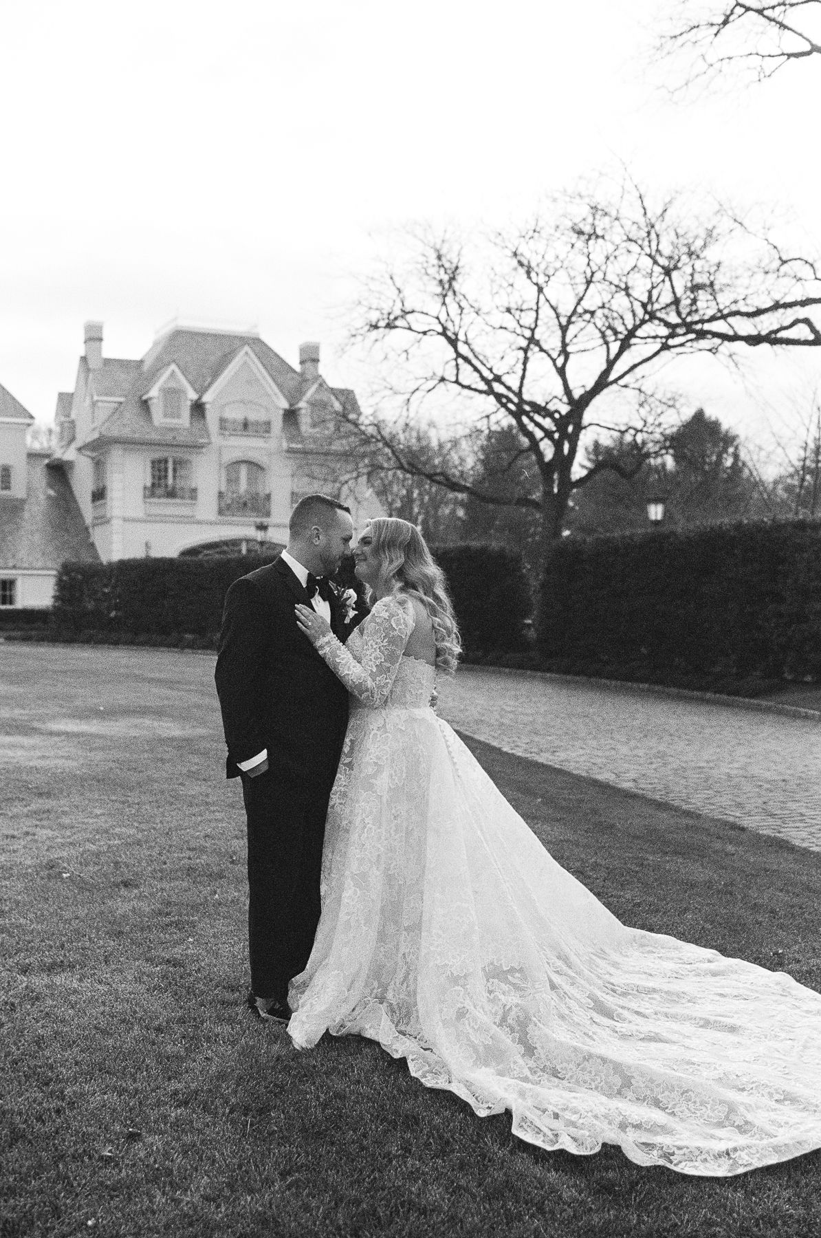Black and white wedding photo of bride in lace gown and groom kissing outdoors near elegant mansion.