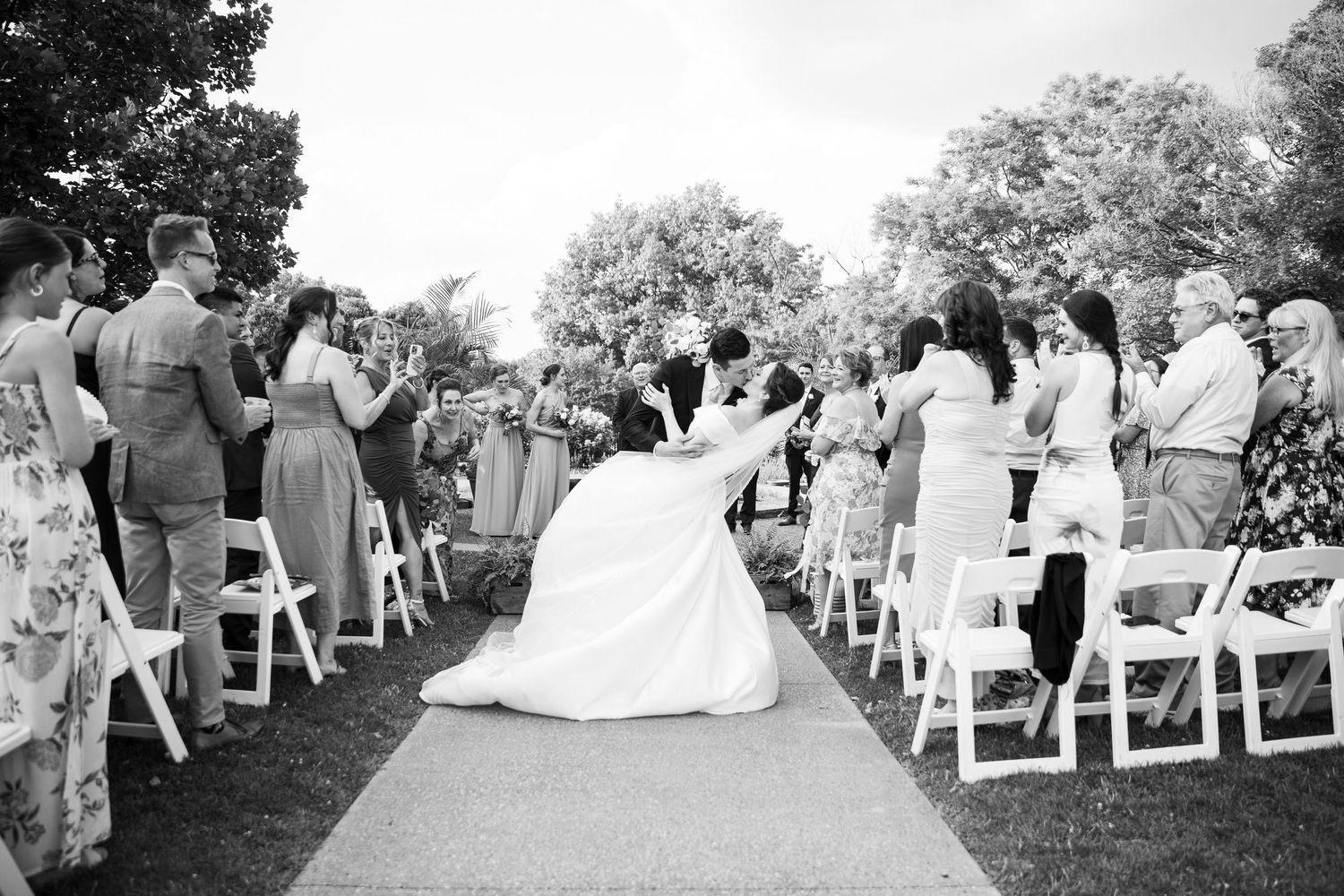 Wedding ceremony guests seated in white chairs on a lawn during an outdoor celebration.