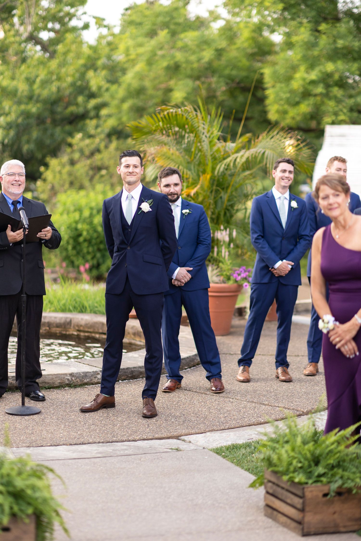 Wedding party members in navy suits and purple dresses pose playfully on a garden path.