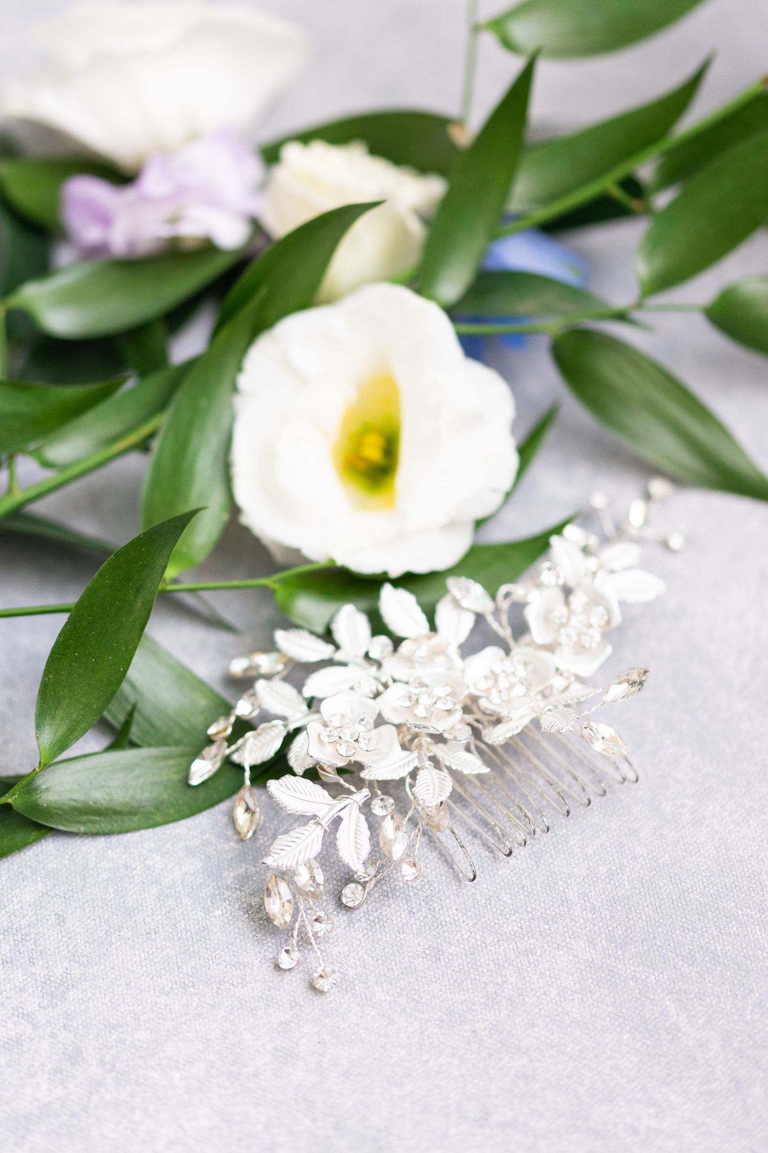 Delicate crystal hair accessories and white flowers arranged with green leaves on a light gray surface.