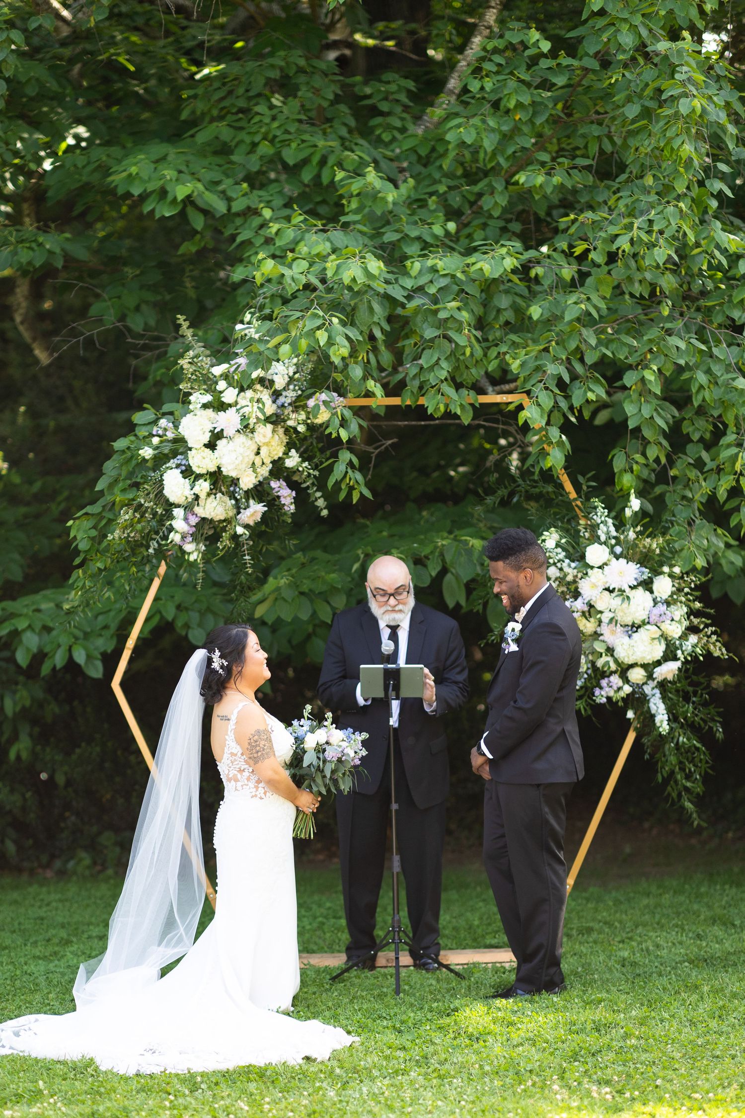 Wedding ceremony in progress under trees with white floral decorations and wooden arch.