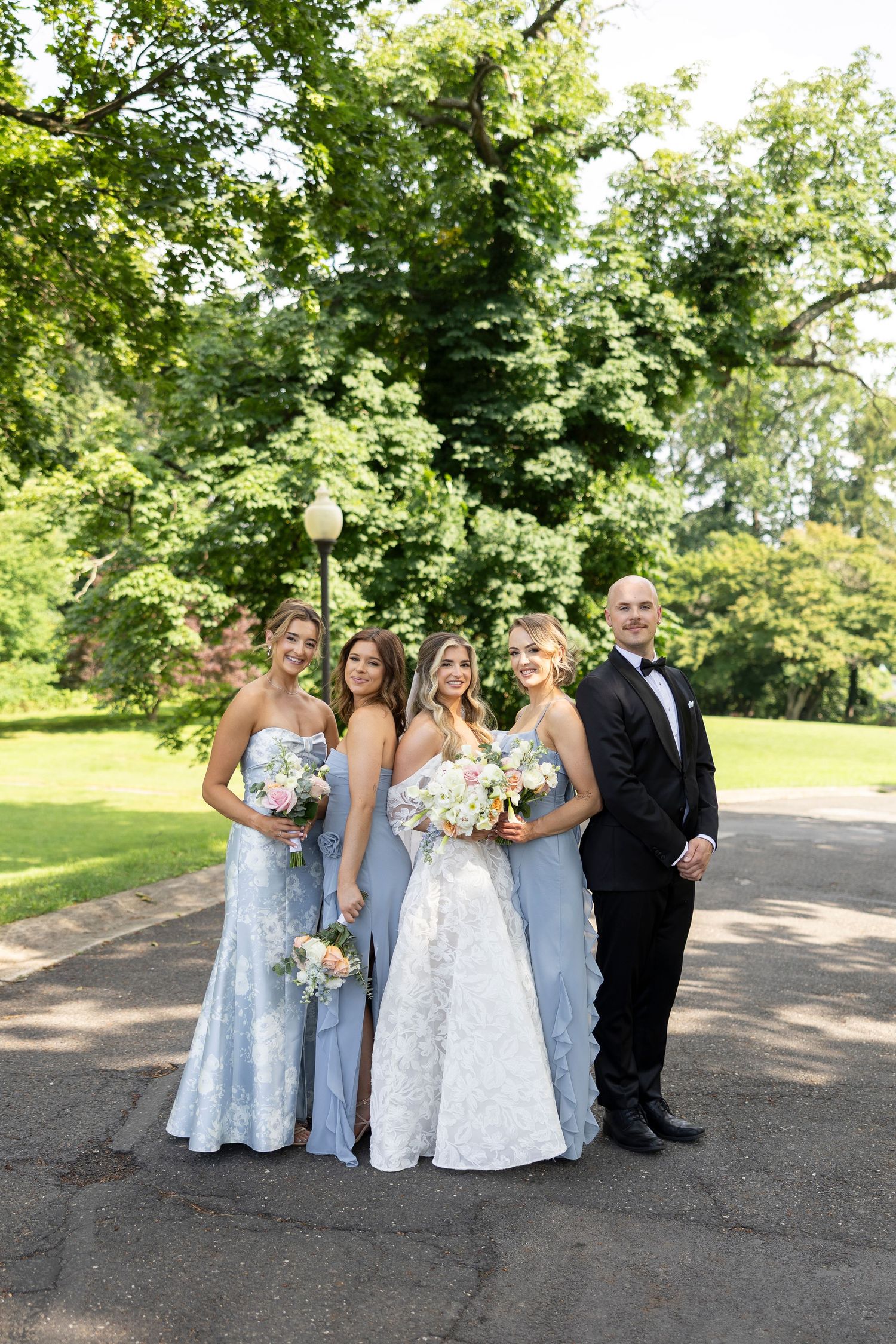 Wedding party in blue dresses and black tuxedo posing together on outdoor path.