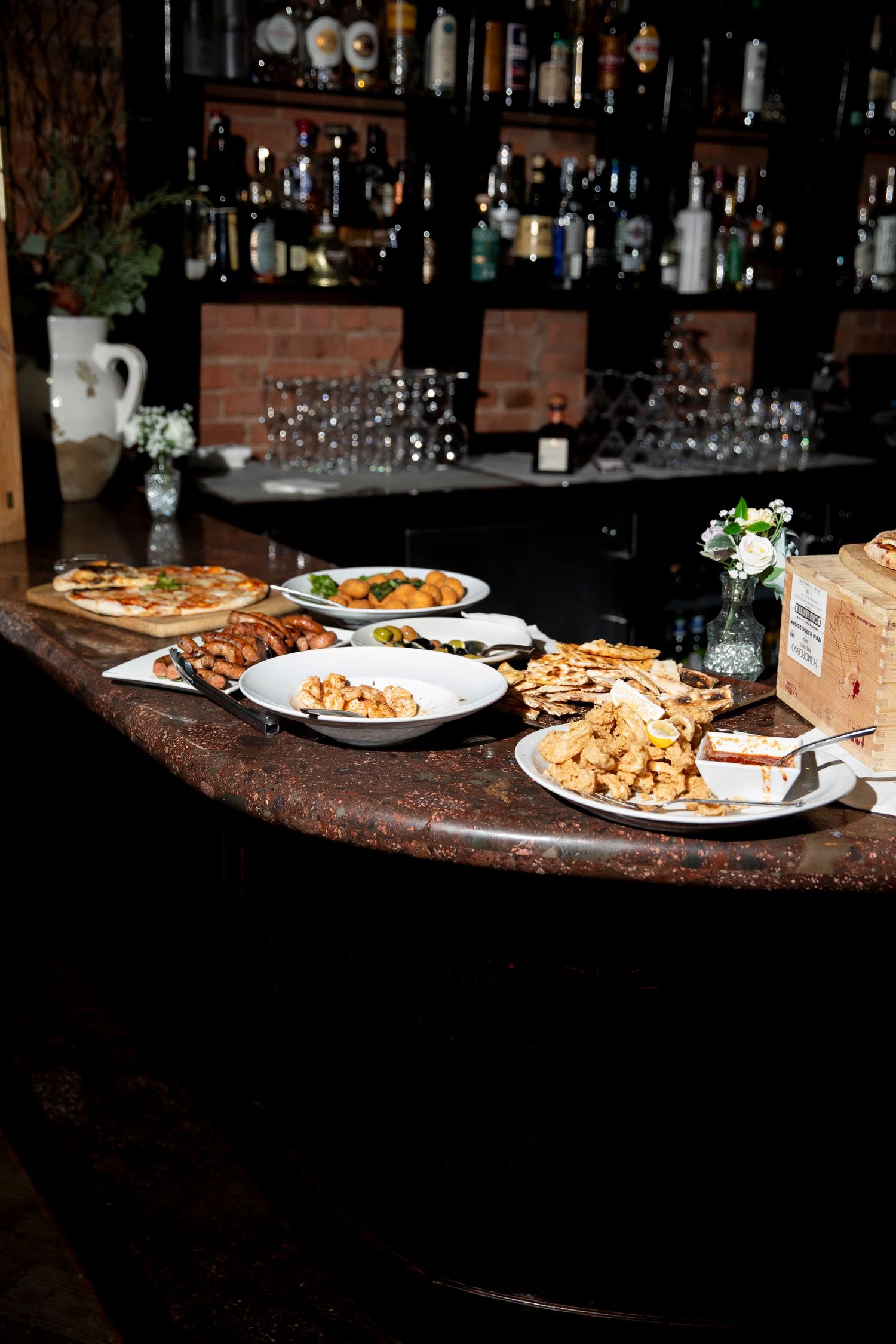 Food plates and drinks arranged on a dark bar counter with bottles in the background.