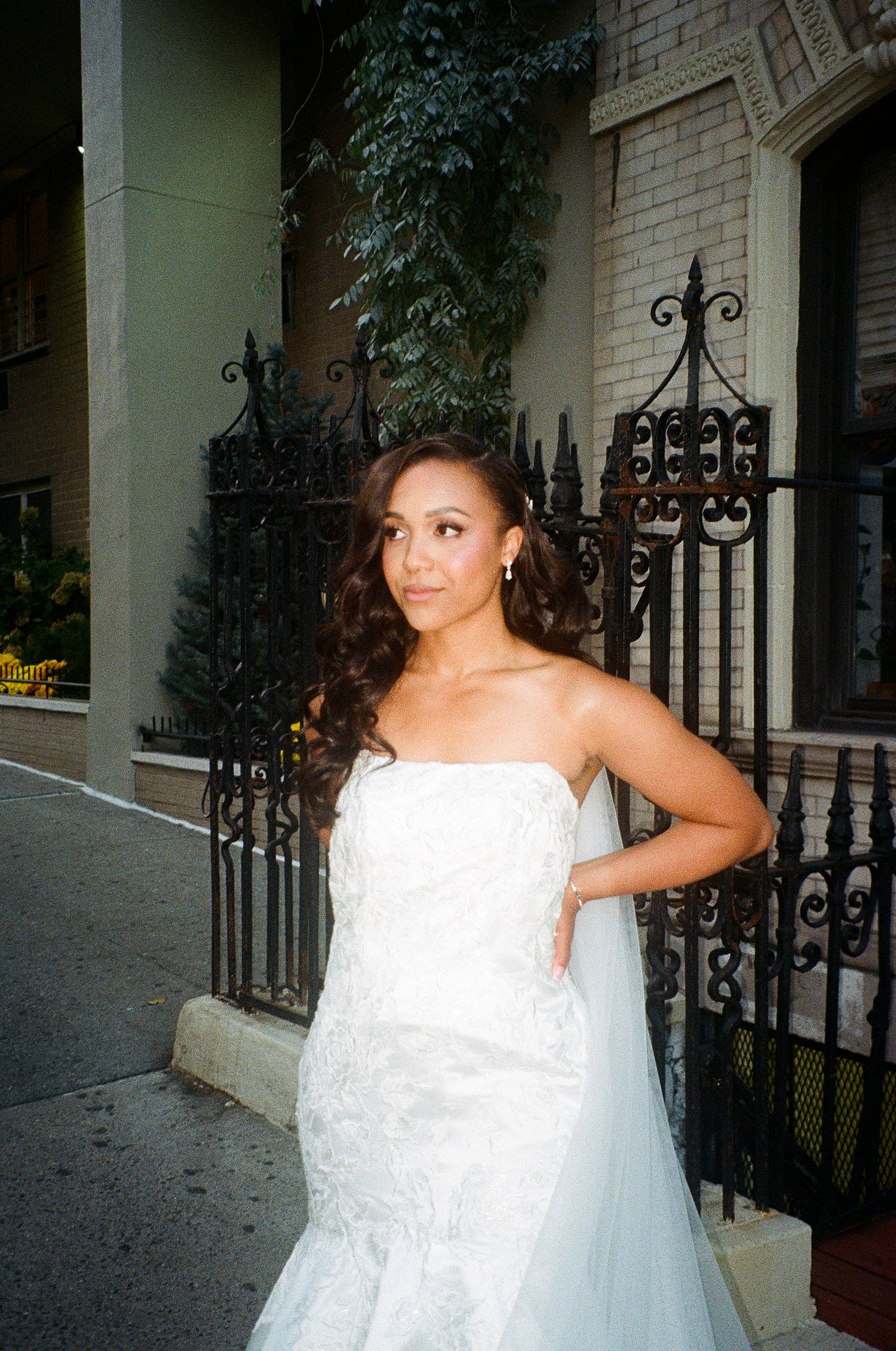 A bride in a flowing white dress stands by decorative ironwork and brick architecture.