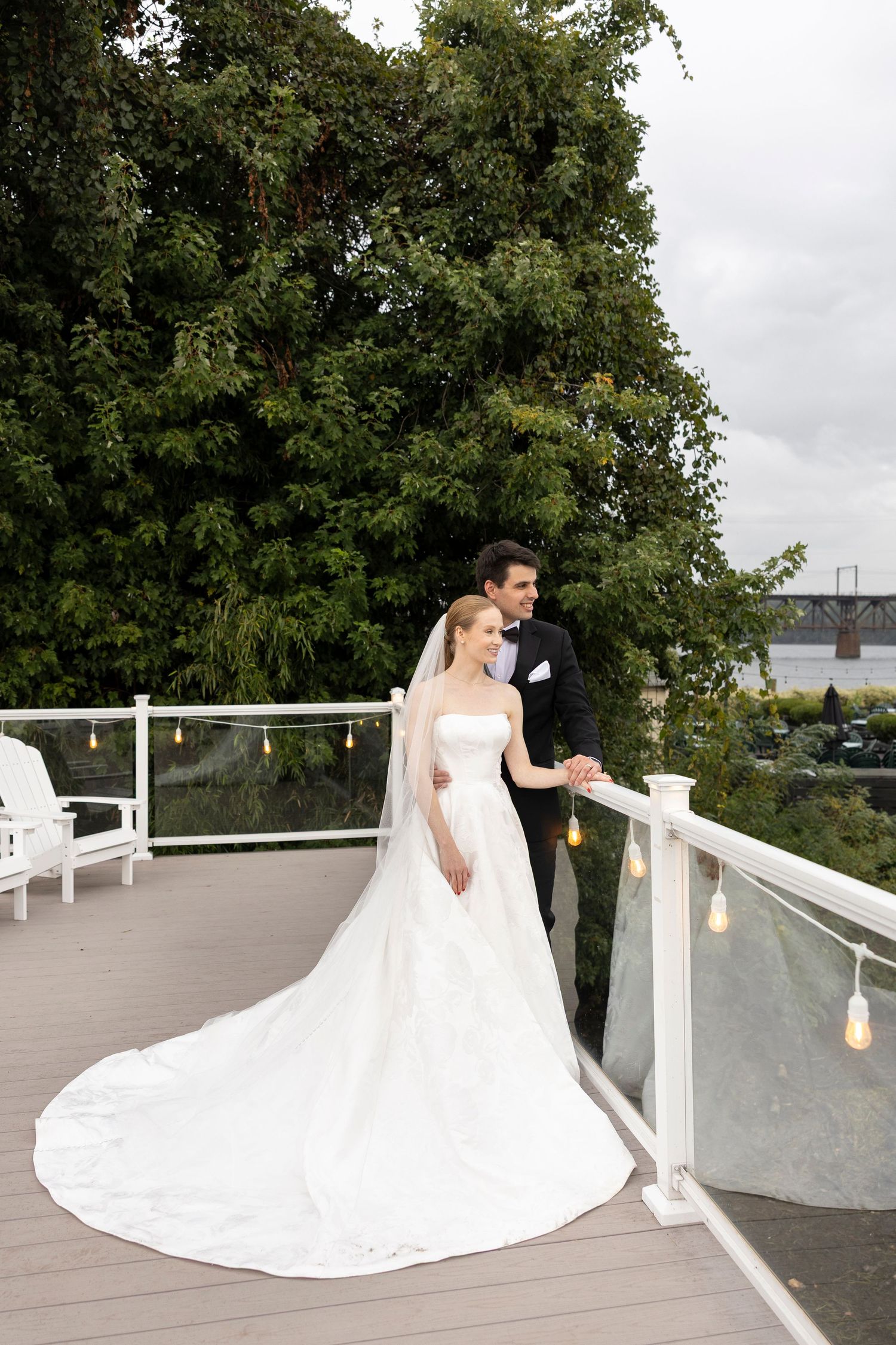 A wedding couple poses on a wooden deck with white railings and lanterns at sunset.