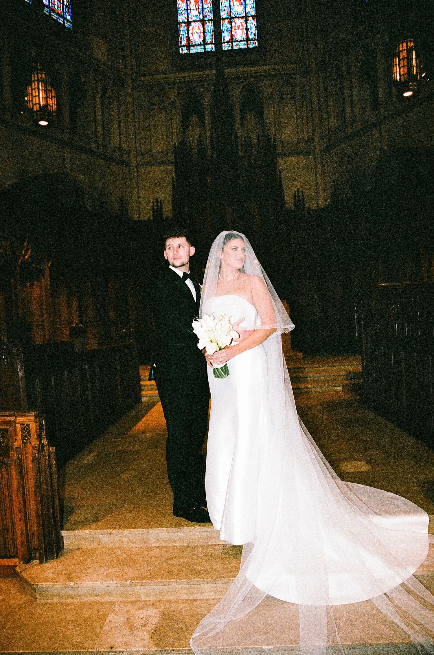 Bride and groom standing at church altar, bride in white gown with long veil holding bouquet, groom in black tuxedo.