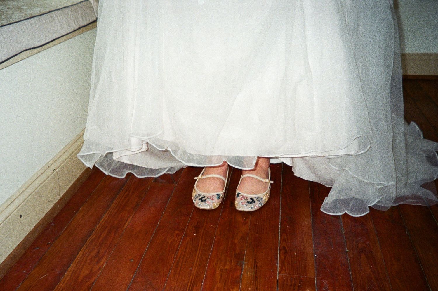Close-up of a bride's white wedding dress hem and decorative flats on a rustic hardwood floor.