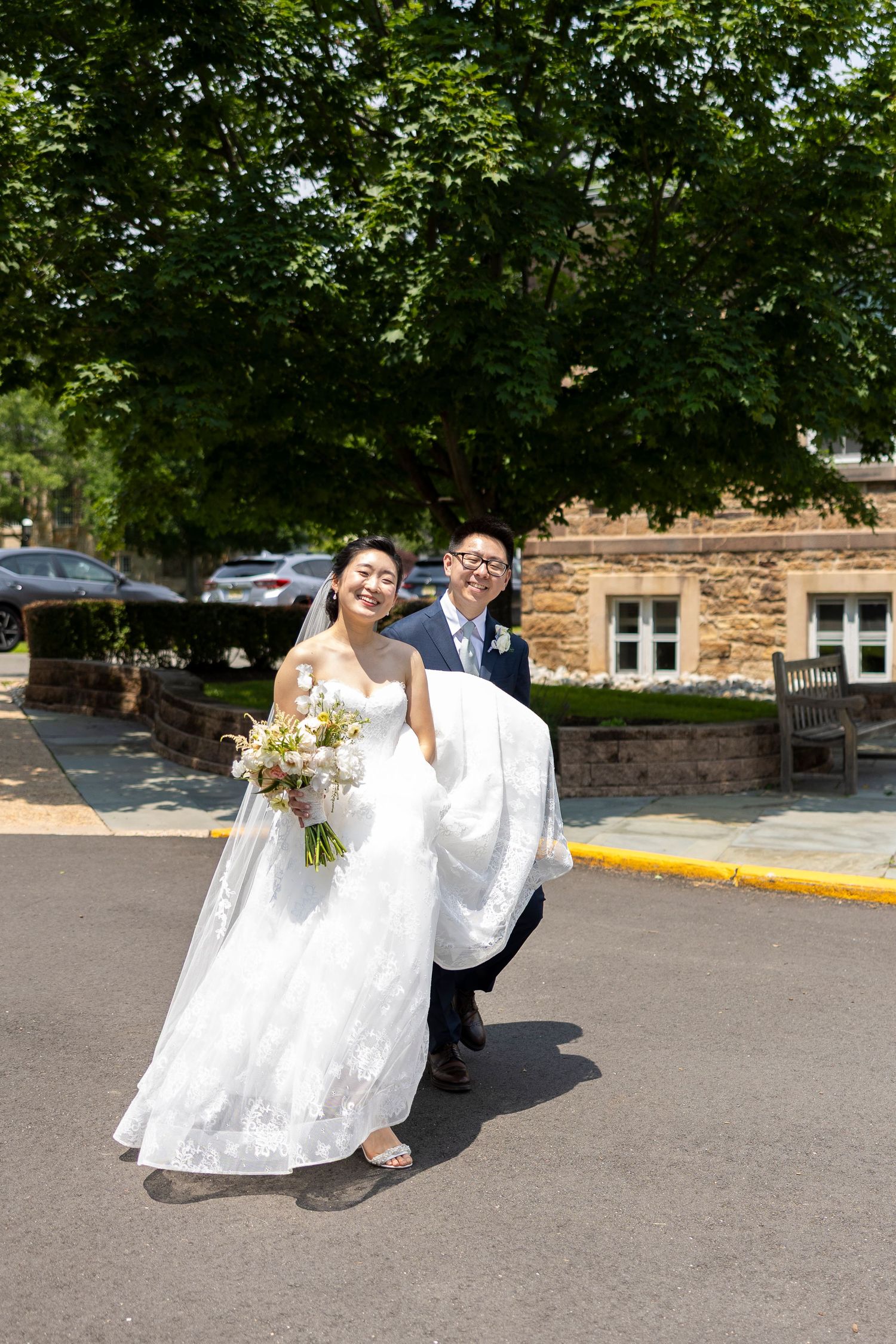 Smiling bride in white gown holding bouquet walks with groom carrying her train outdoors on sunny wedding day.