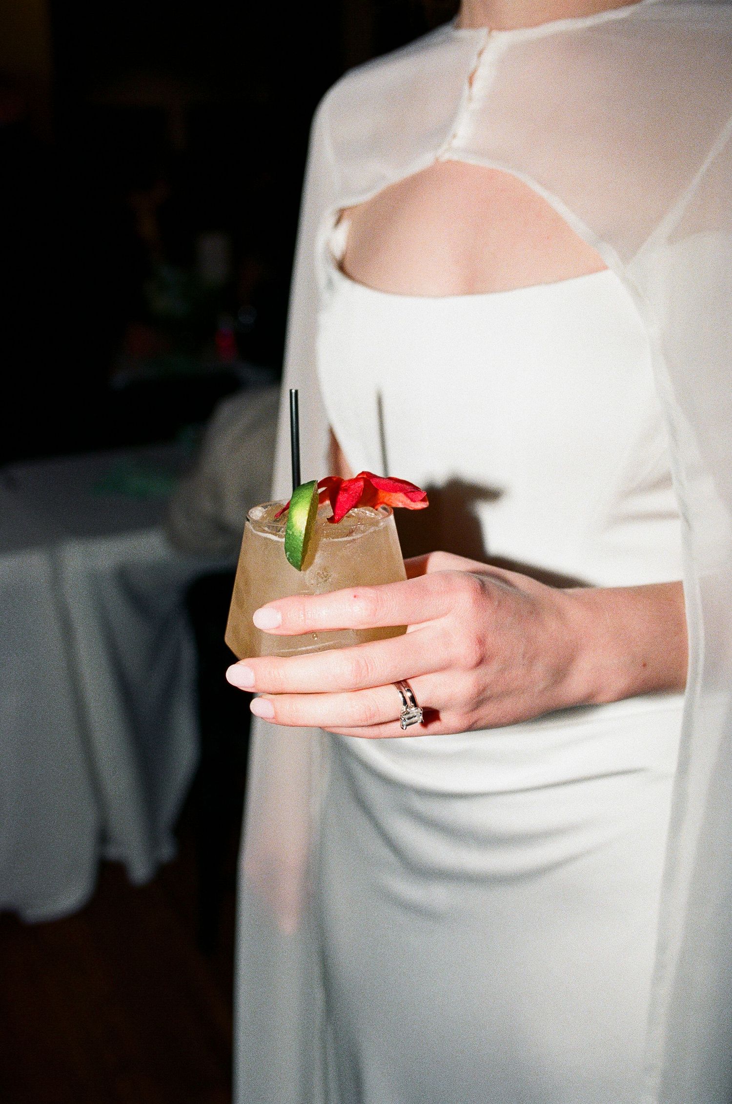 Bride in white cutout dress holding cocktail with red flower garnish and engagement ring visible.