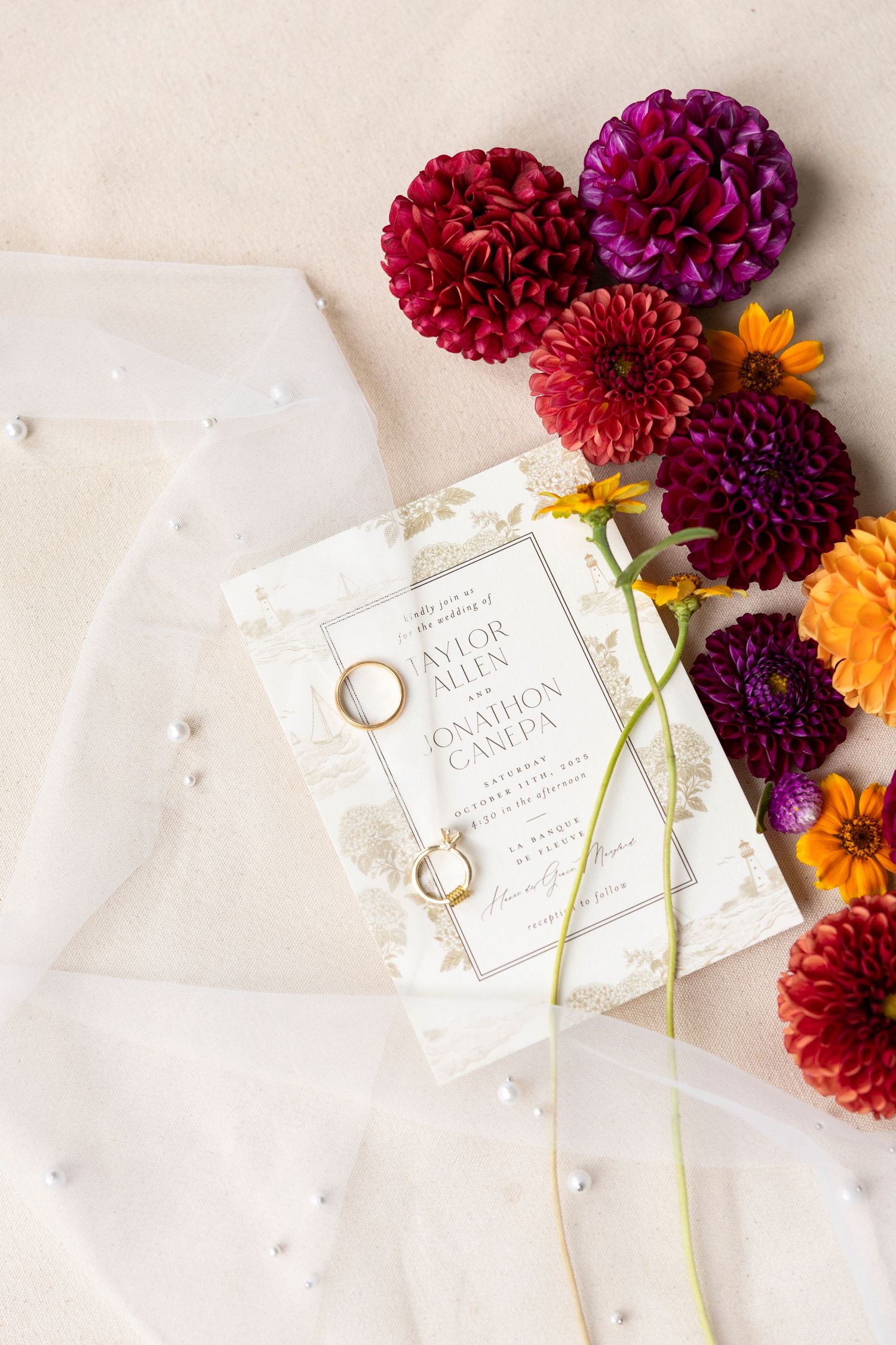 Wedding invitation and rings styled with burgundy and orange dahlias on white surface.