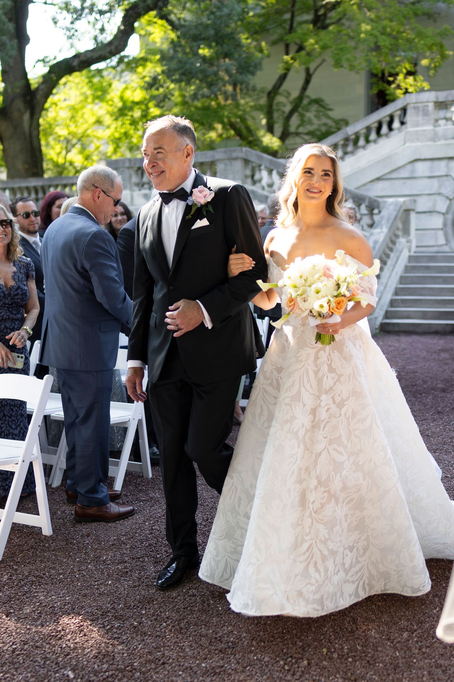 Wedding ceremony attendees gather on a gravel path surrounded by trees and stone architecture.