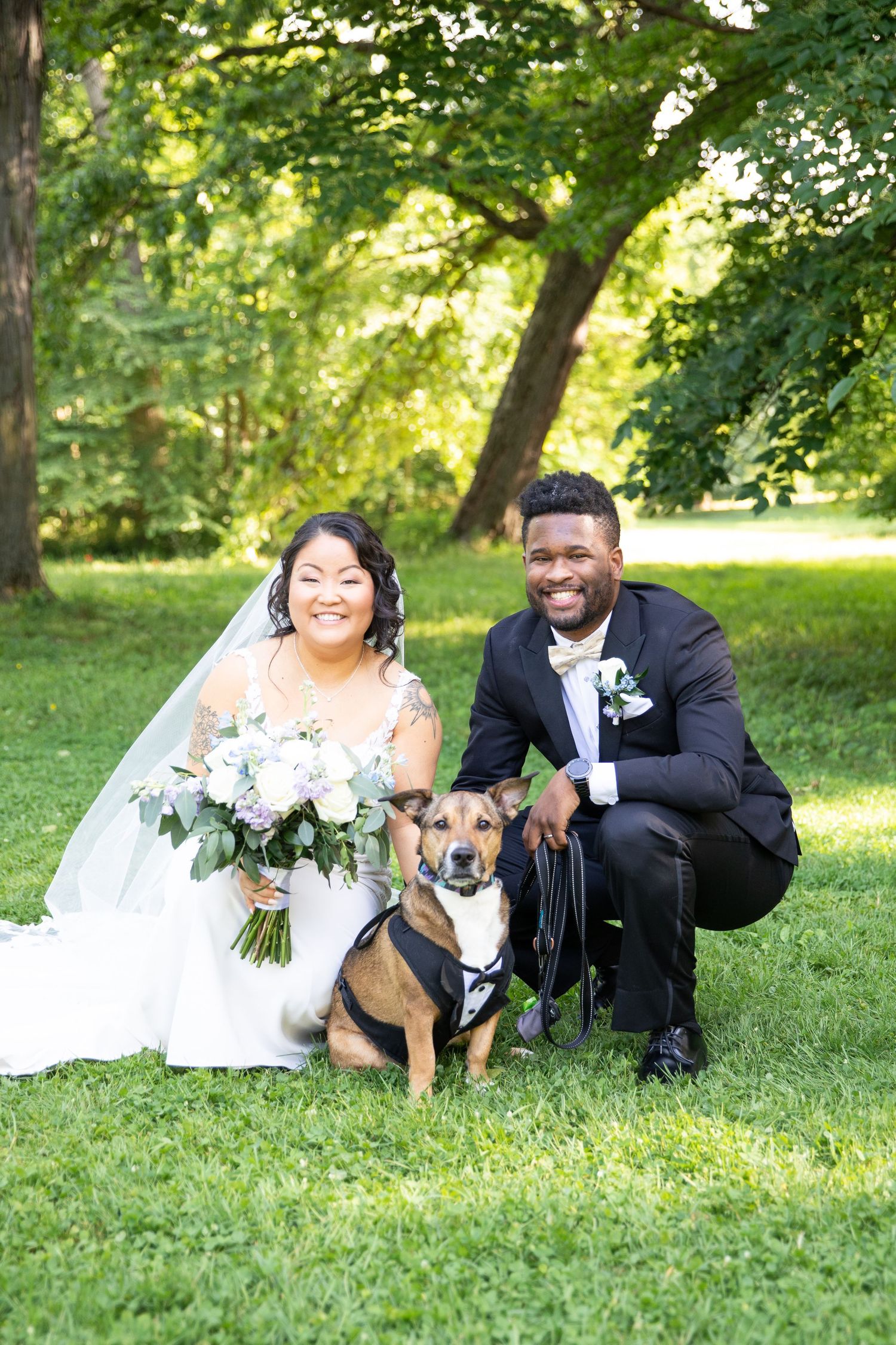 A couple in wedding attire poses on grass with their dog during an outdoor photo session.