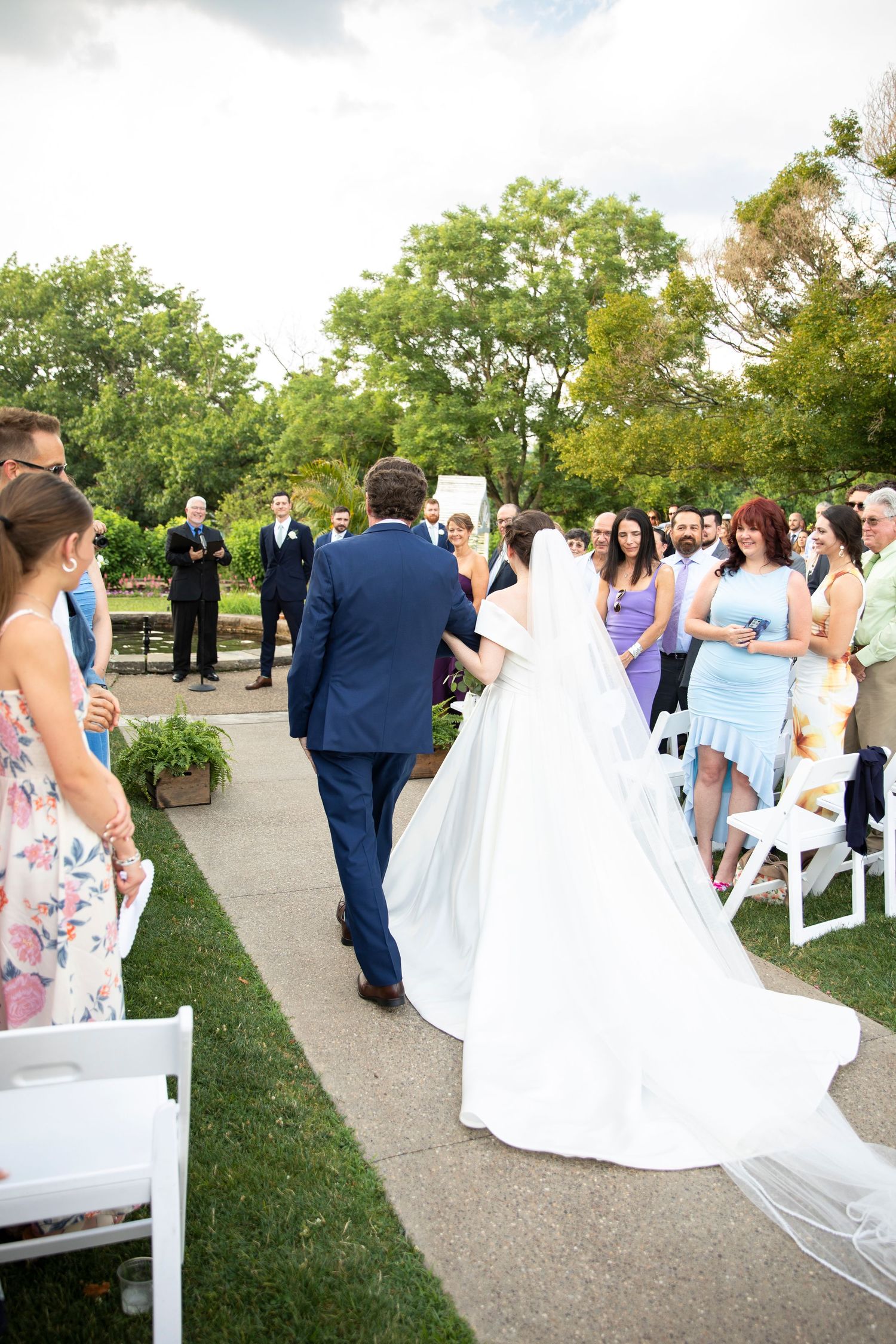 Outdoor wedding ceremony with guests seated on white chairs as couple exchanges vows on stone pathway.