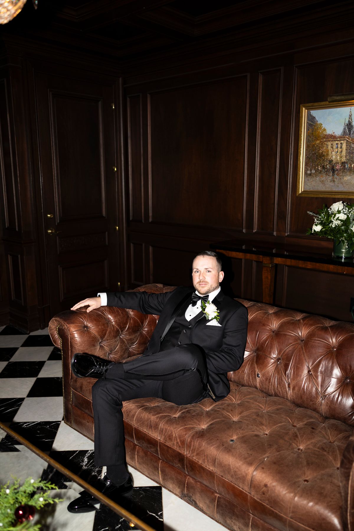 Groom in black tuxedo with white boutonniere relaxing on tufted brown leather Chesterfield sofa in elegant wood-paneled room.