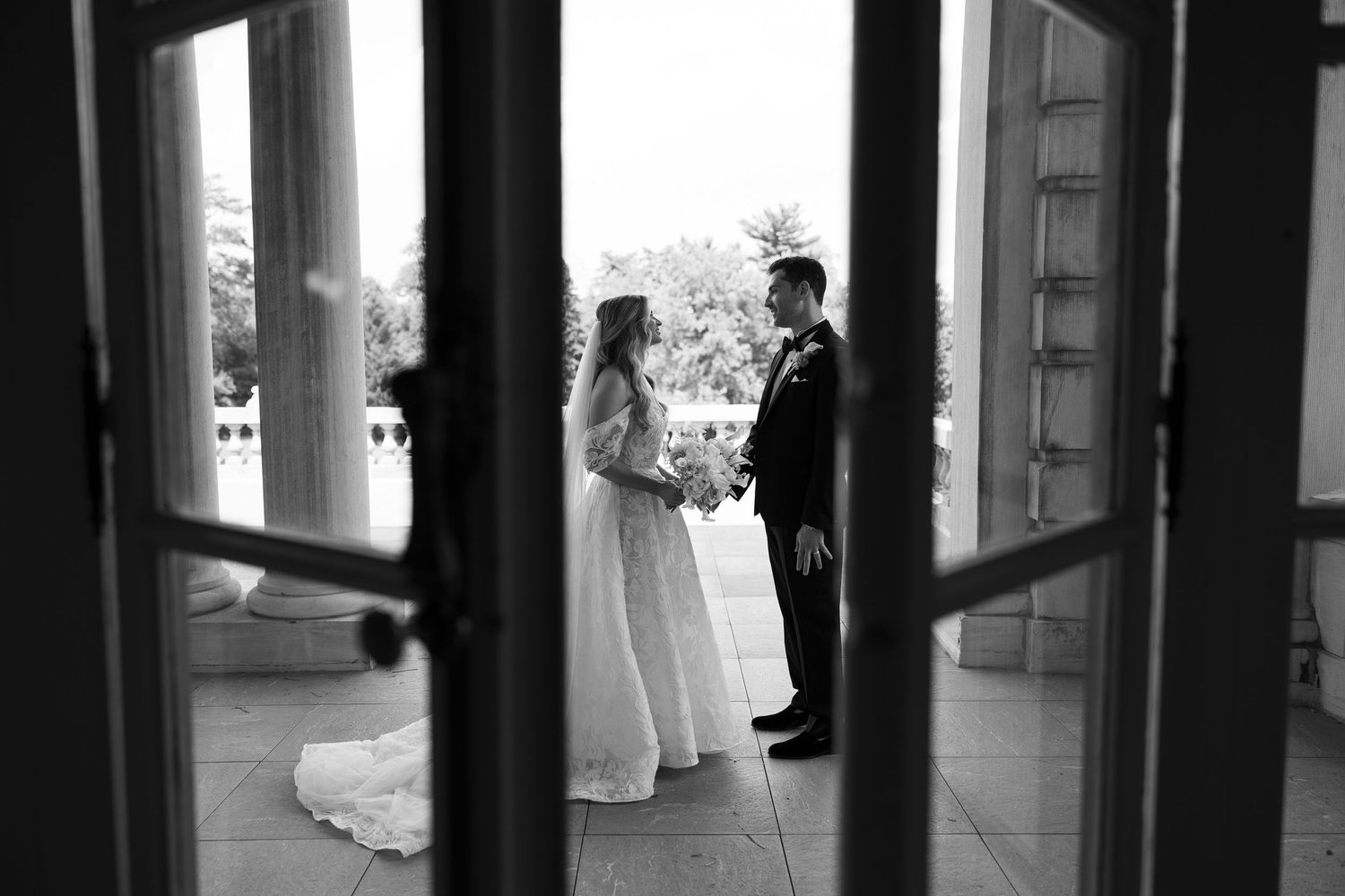 Black and white photo of bride and groom exchanging vows framed by elegant open doors at a classic wedding venue.