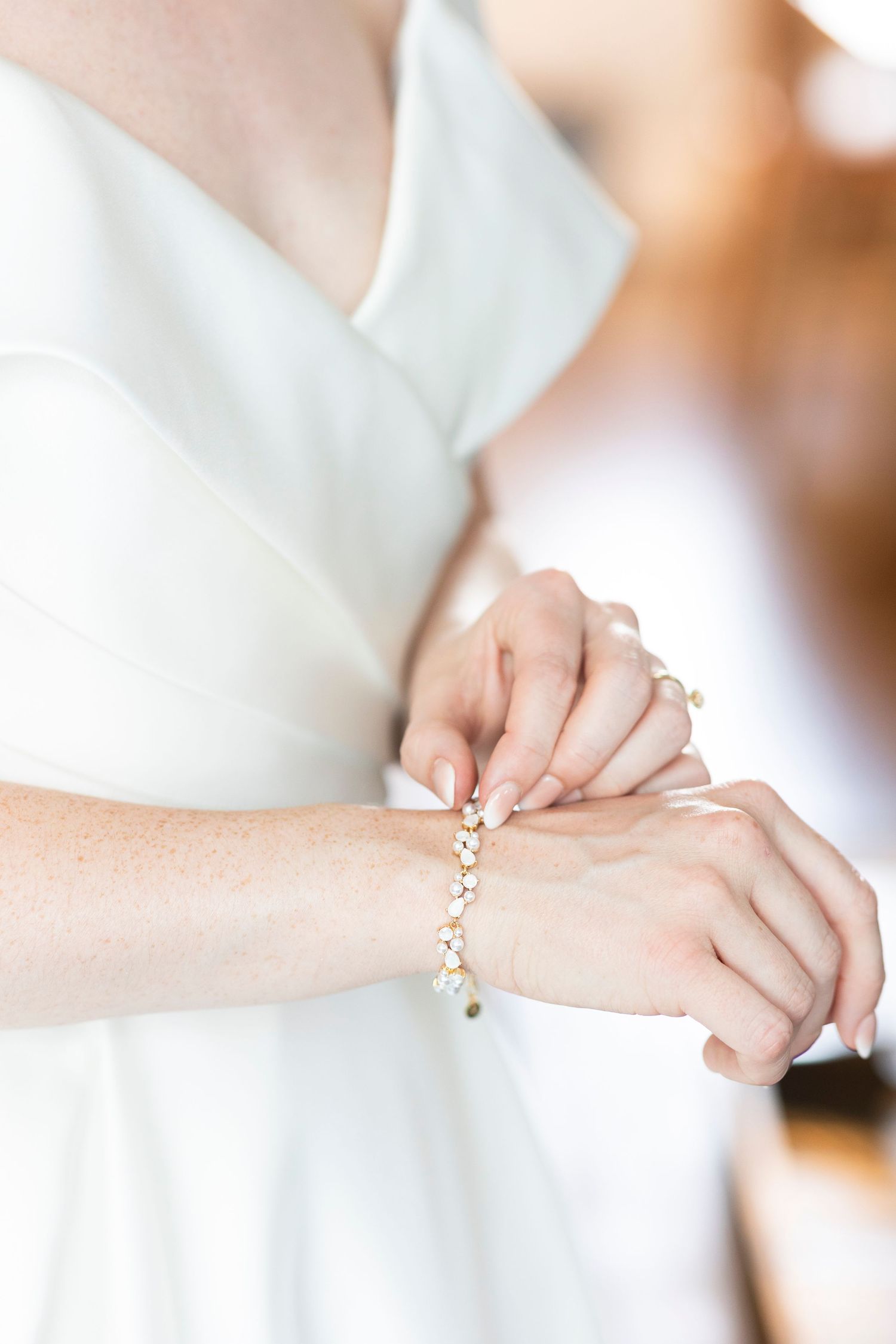 Close-up of hands fastening an elegant pearl bracelet against a white wedding dress.