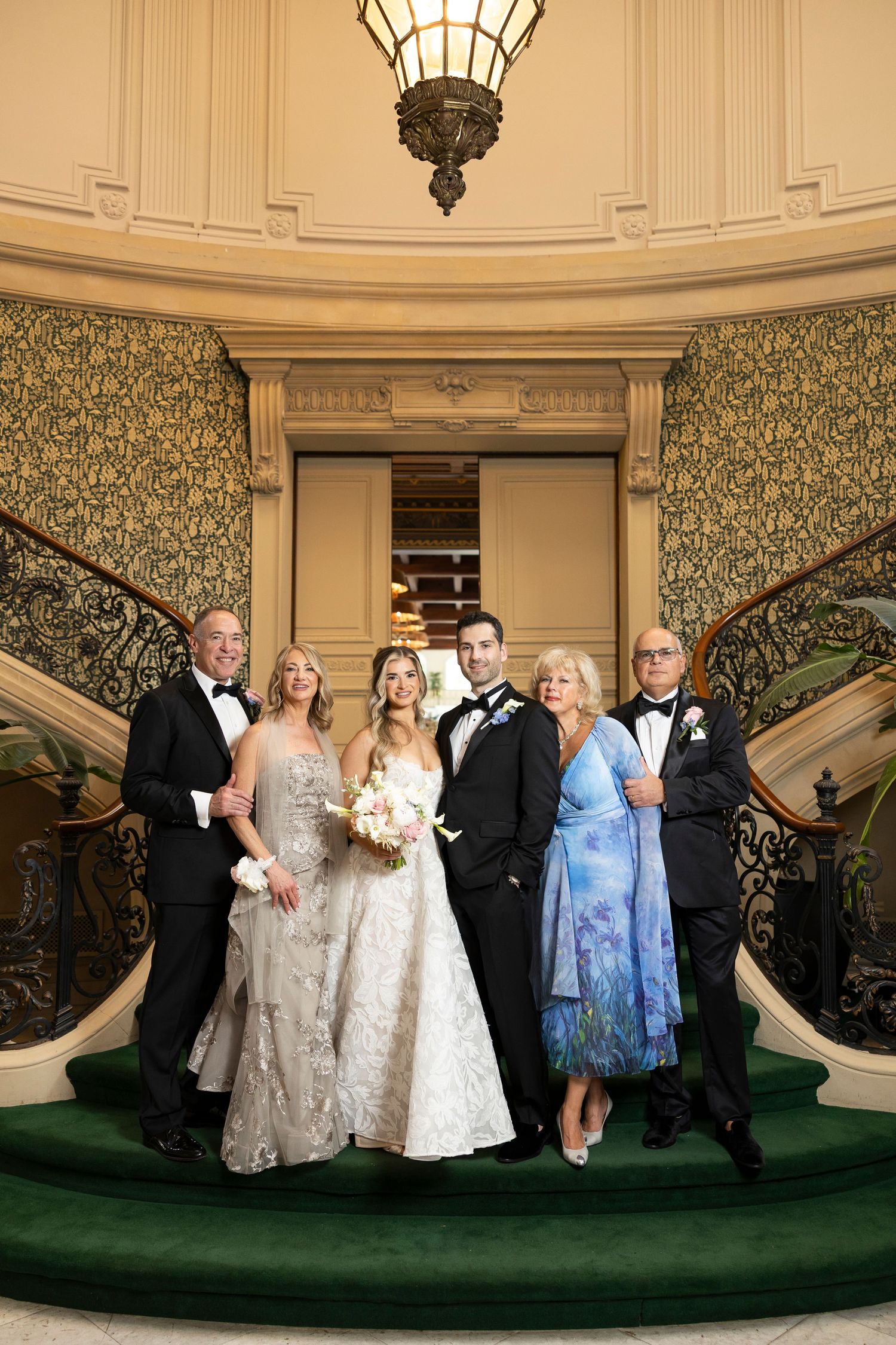 Wedding party gathered on elegant staircase with green carpet and decorative wallpaper.