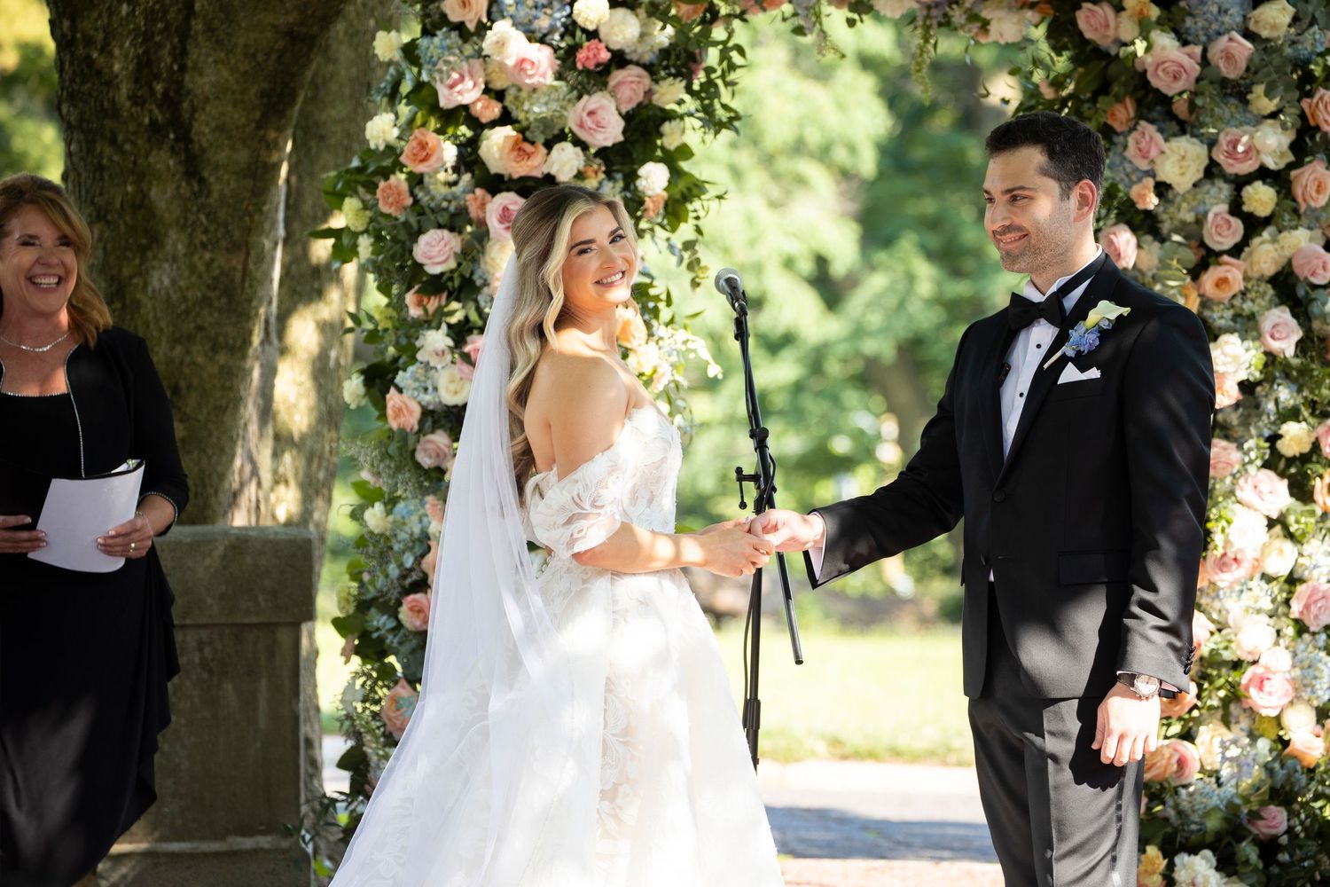 Wedding ceremony under a floral arch with pink and peach blooms in an outdoor garden setting.