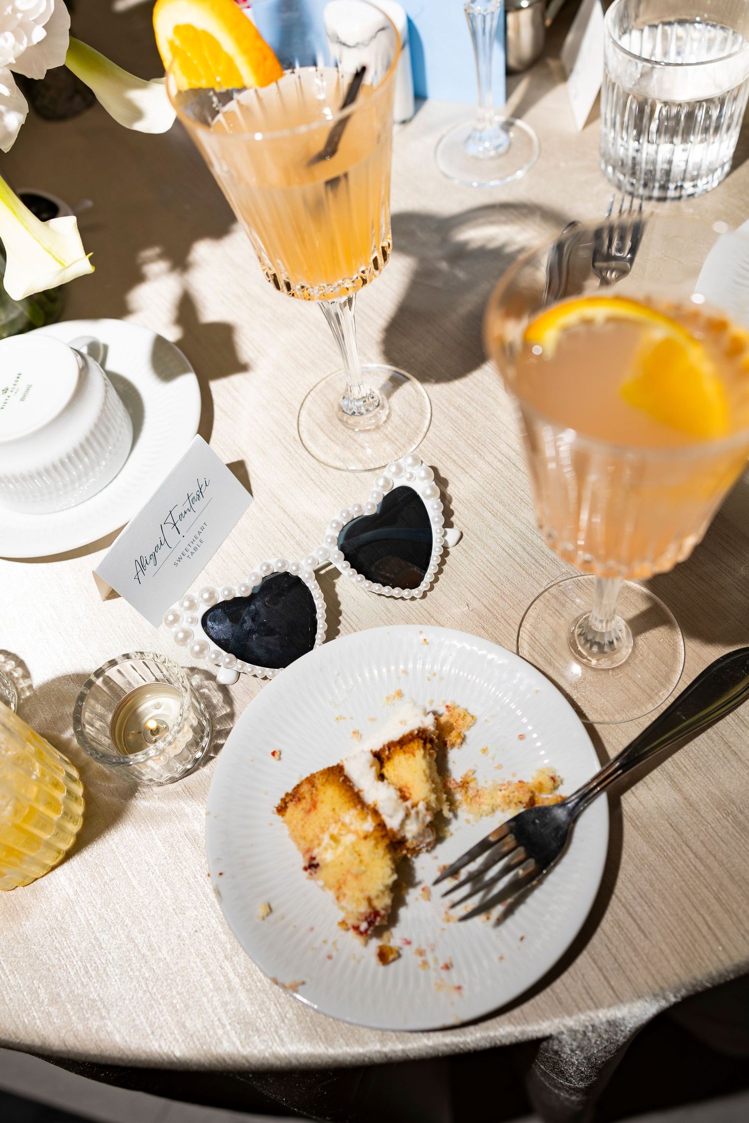 Half-eaten slice of wedding cake with cocktails on white tablecloth.