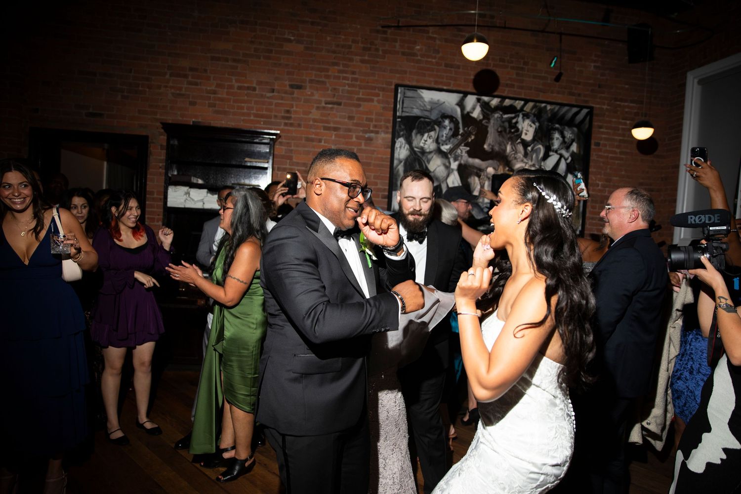 Energetic wedding party dancing and celebrating in a dimly lit venue with brick walls.