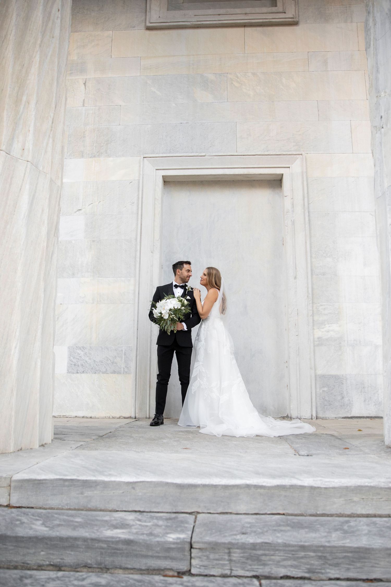 Bride and groom posing elegantly in front of a marble wall, bride holding white bouquet in a lace gown.