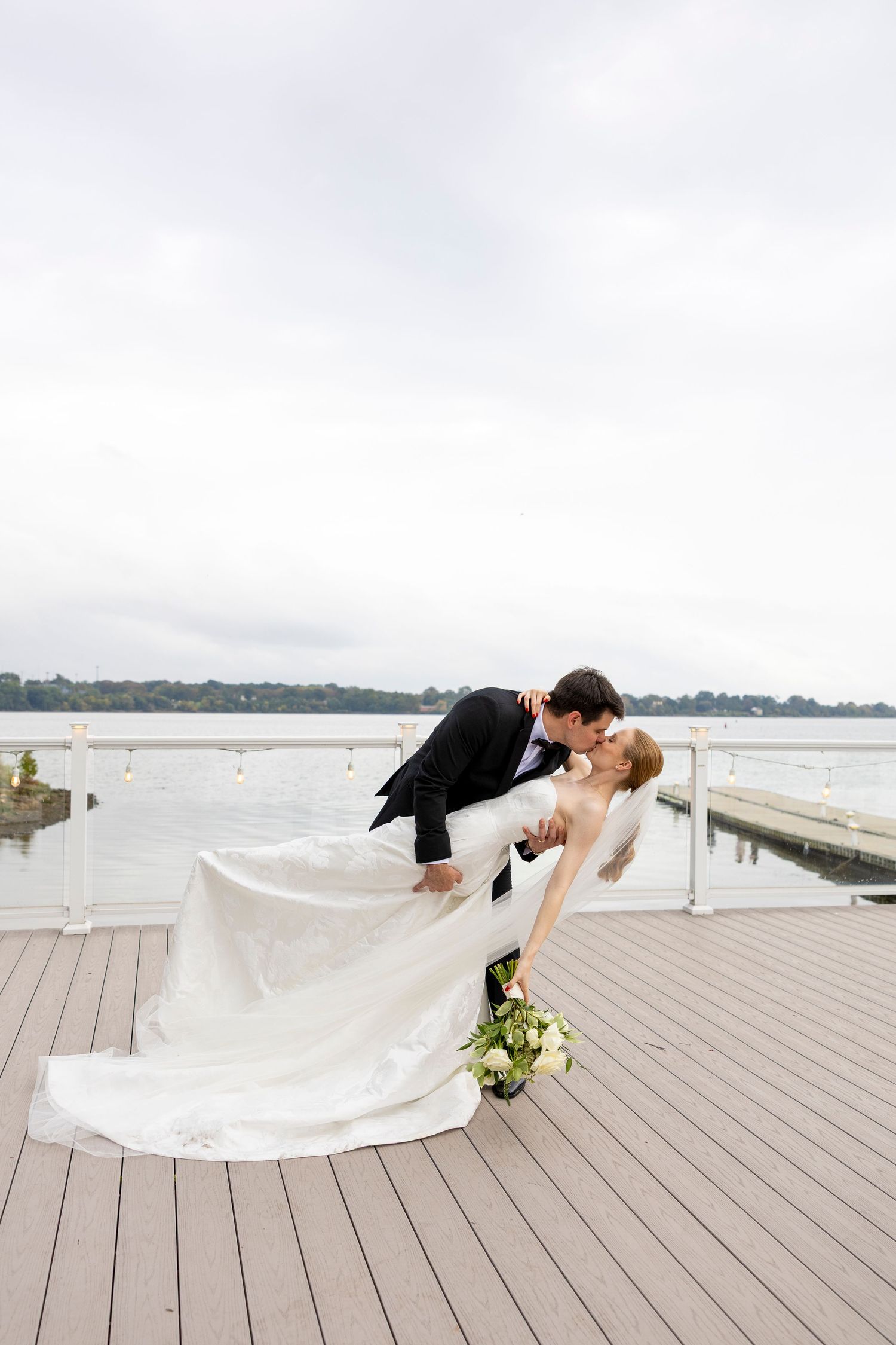 Groom dips bride for a romantic kiss on a waterfront deck, her white gown flowing, bouquet in hand, under a cloudy sky.