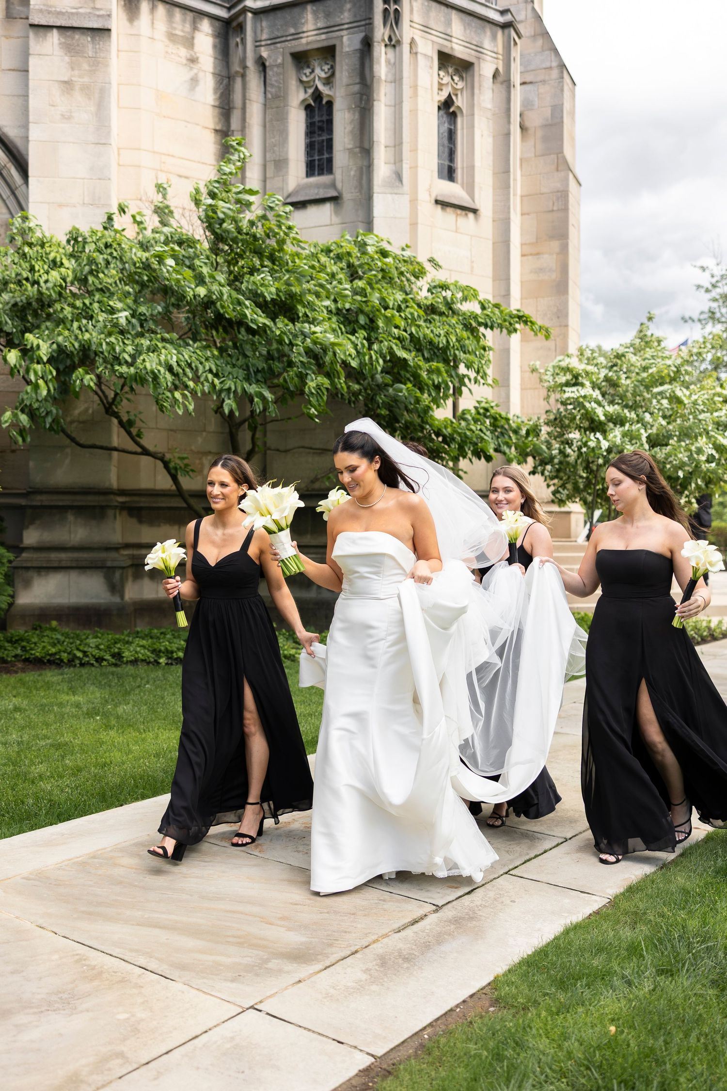 Bride in white gown walks with bridesmaids in black dresses, holding bouquets outside a stone church.