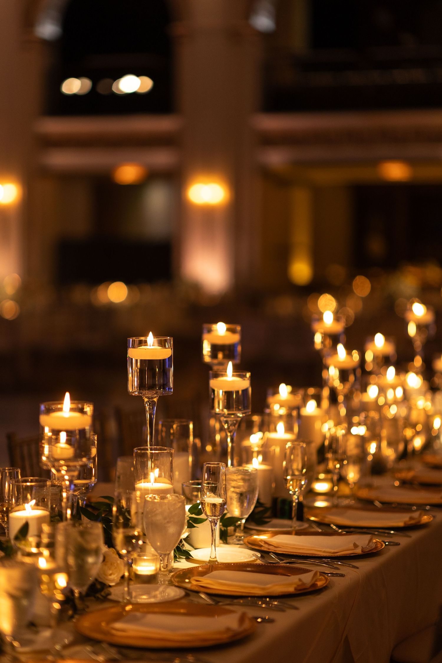 Elegant candlelit reception table with floating candles in glass holders, gold charger plates, and crystal glassware.