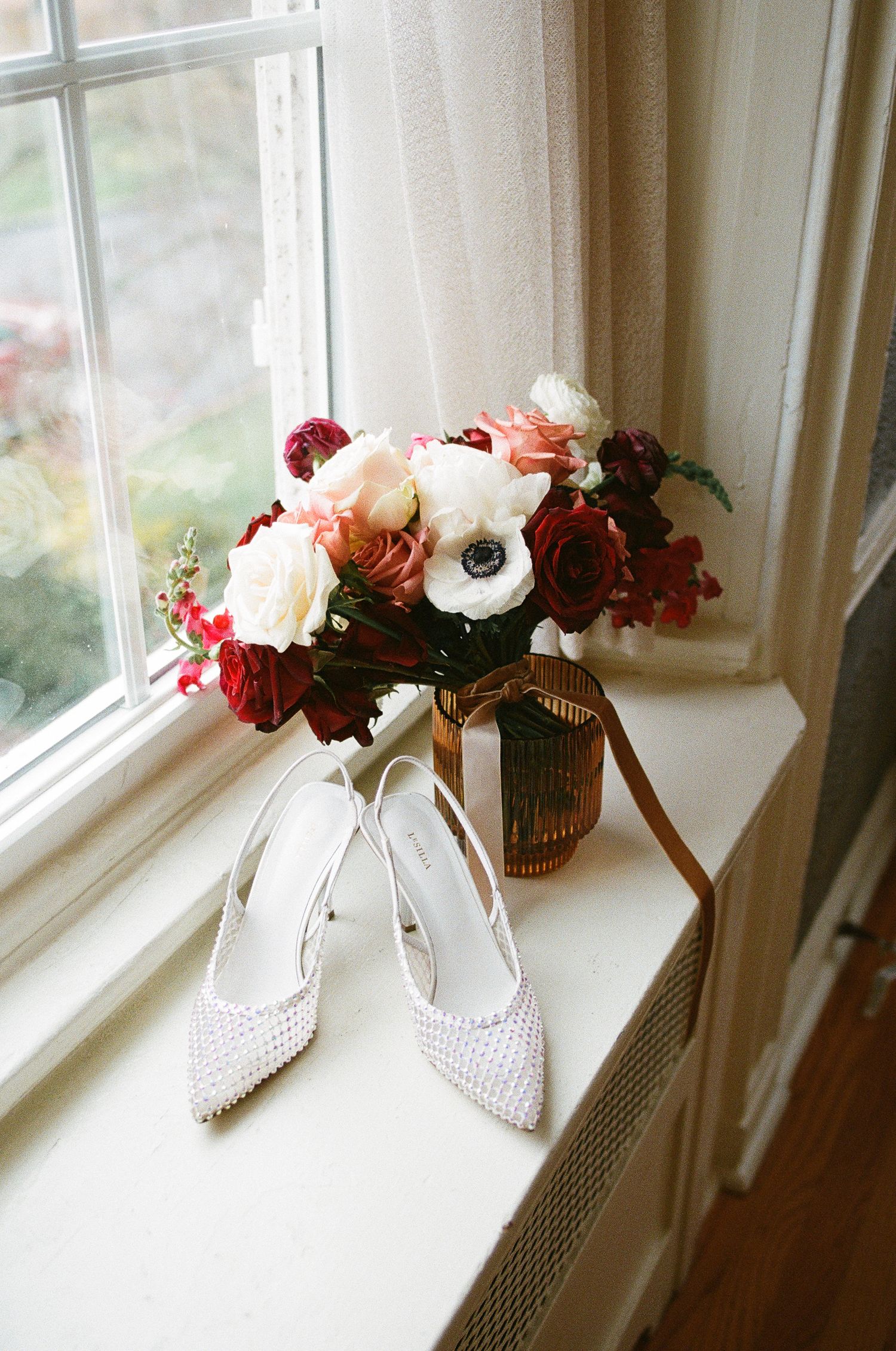 Pearl bridal heels leaning against a gold vase with red, pink, and white wedding bouquet on a sunlit windowsill.