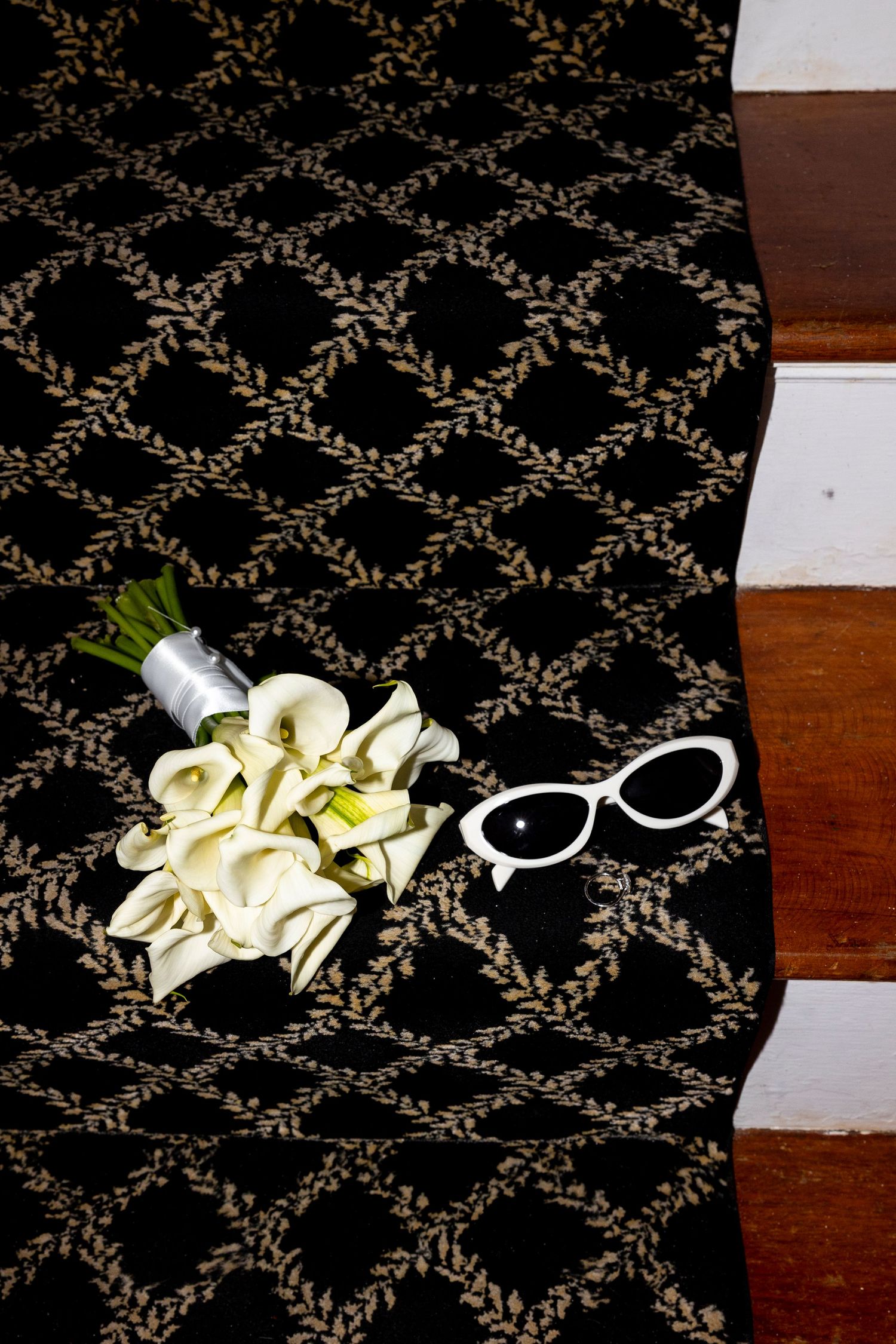 White calla lily bouquet and wedding accessories arranged on dark patterned carpet.