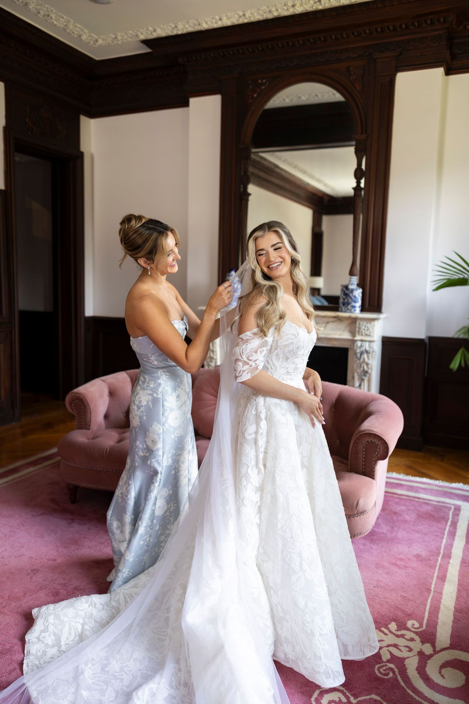 Two women in white lace wedding gowns embrace on a pink carpeted floor.