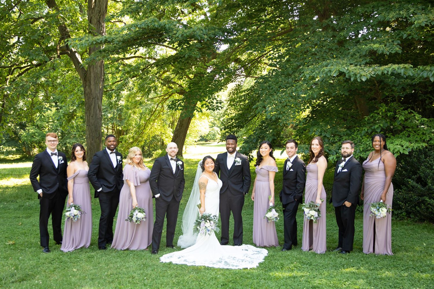 Wedding party arranged in a line outdoors with bridesmaids in lavender dresses and groomsmen in black suits.