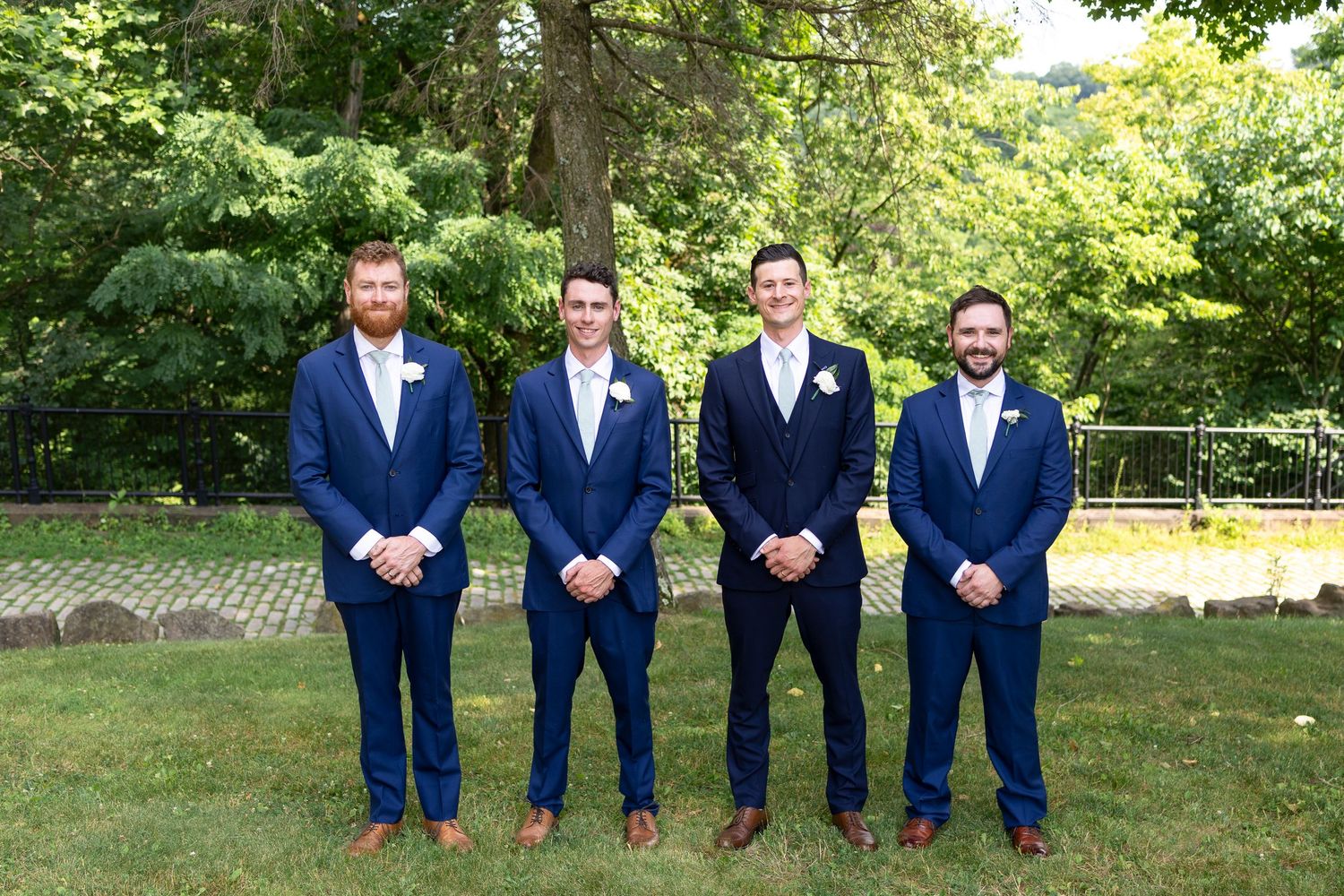 Groomsmen in matching navy suits stand in line on grass under trees.