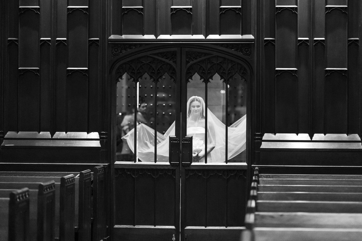 Black and white photo of a bride in veil seen through ornate church doors with wooden pews in foreground.