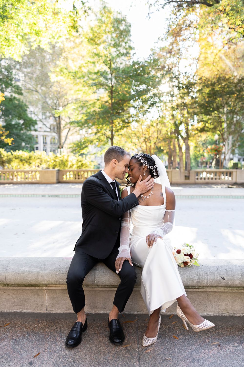 Interracial couple sharing a romantic moment in wedding attire seated by a fountain surrounded by lush green trees.