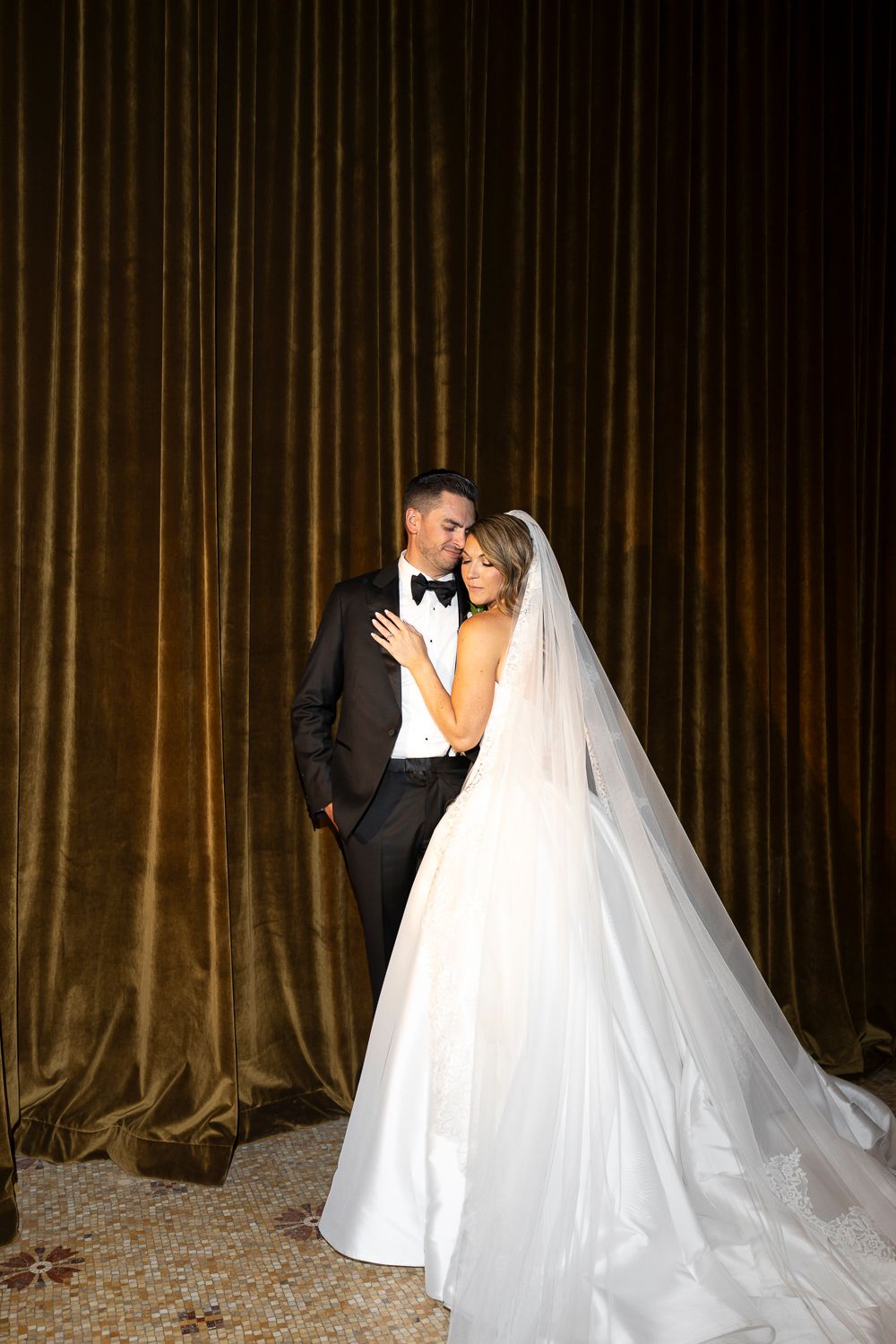Bride in a white ballgown and veil embraces groom in black tuxedo against golden velvet curtains.