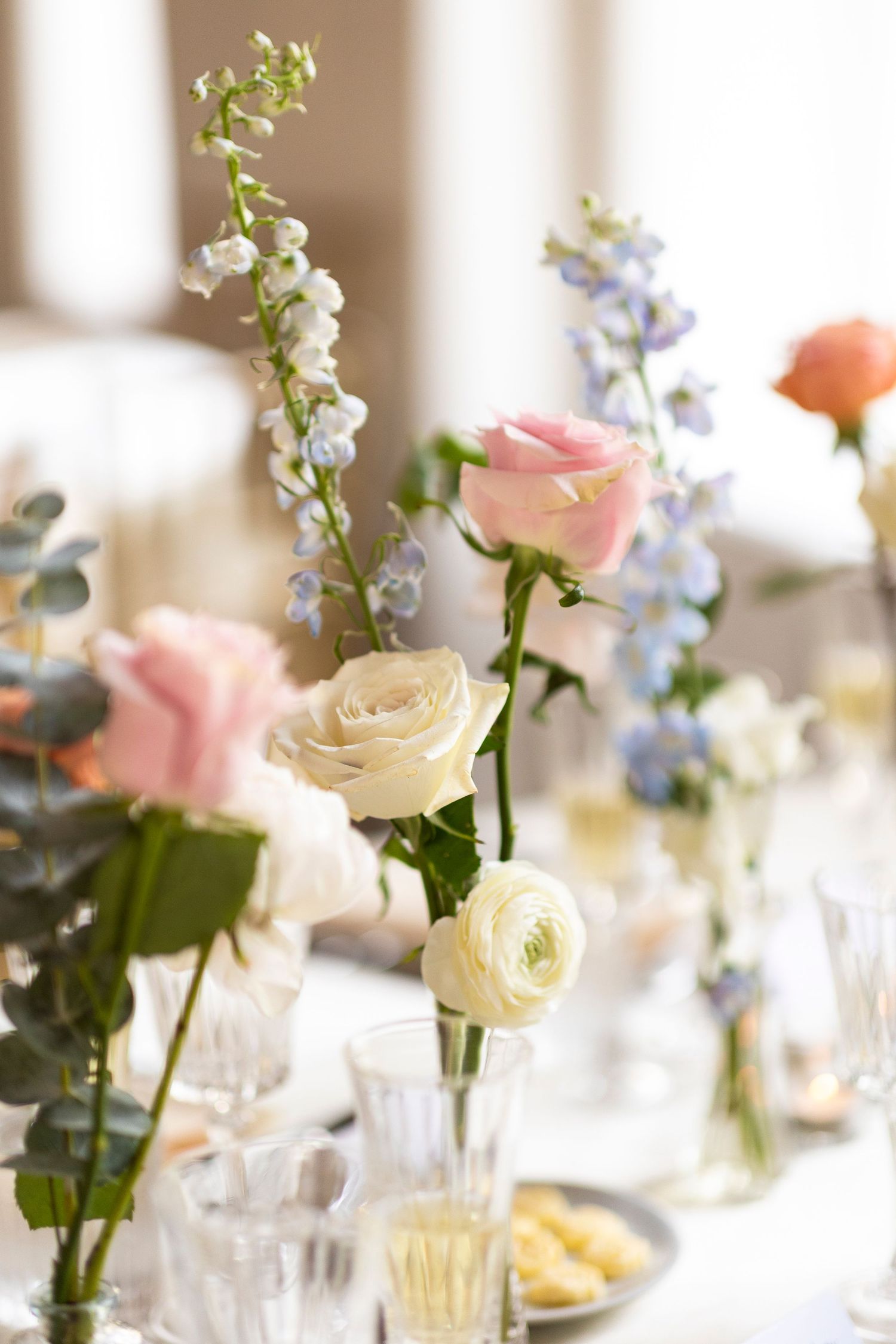 Elegant floral centerpieces with pink roses and white ranunculus in glass vases on a wedding reception table.