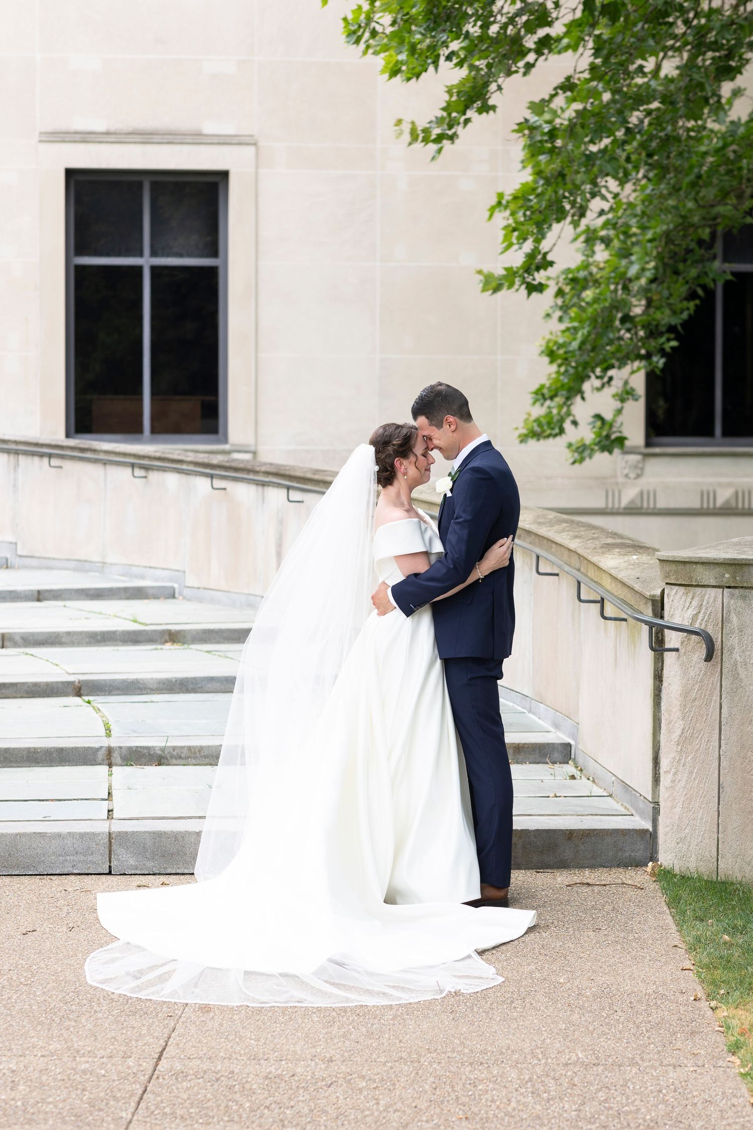Romantic wedding moment captured on stone steps with white building and greenery in background.