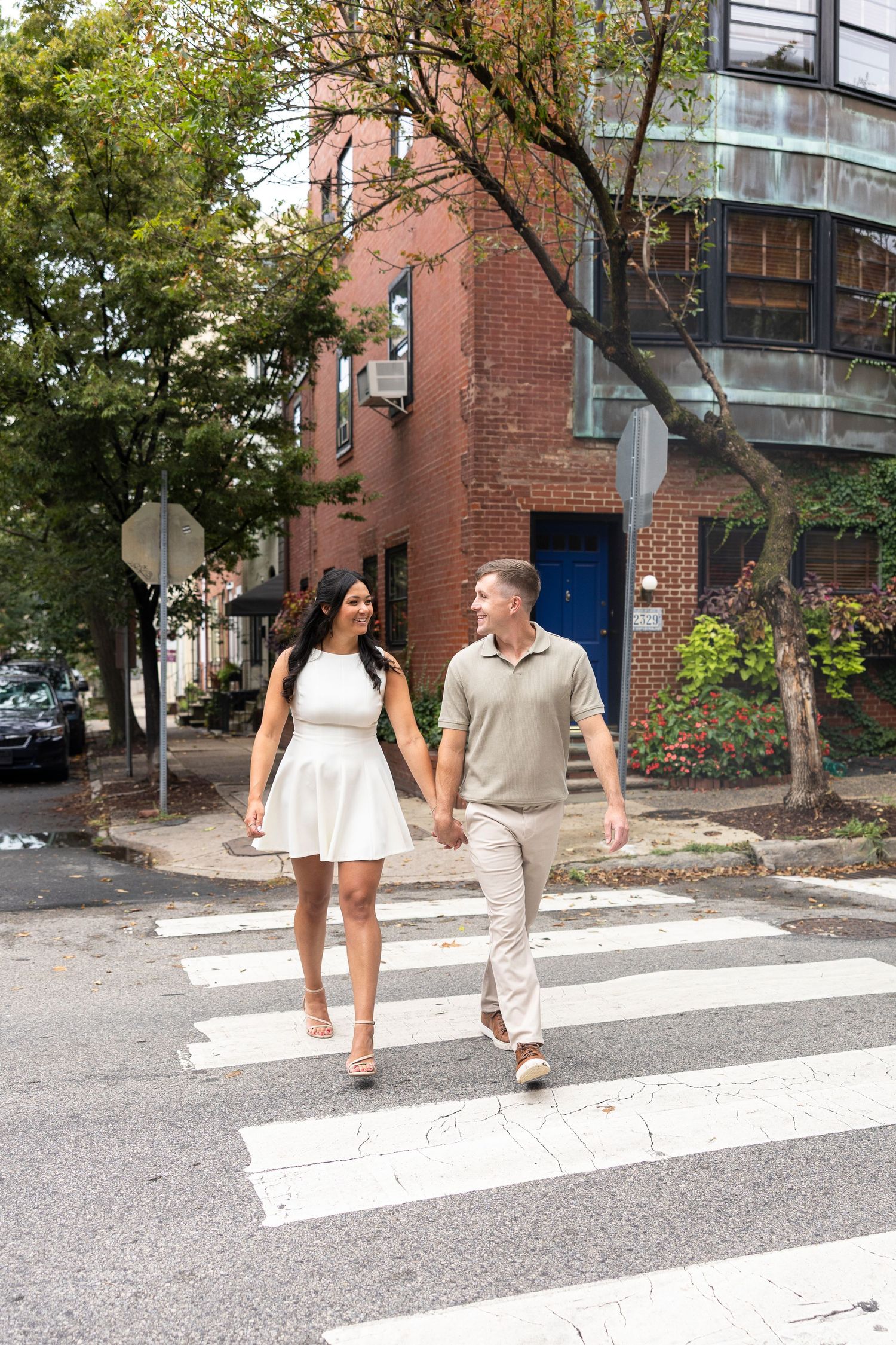 A couple walks across a crosswalk near brick buildings with blue doors.