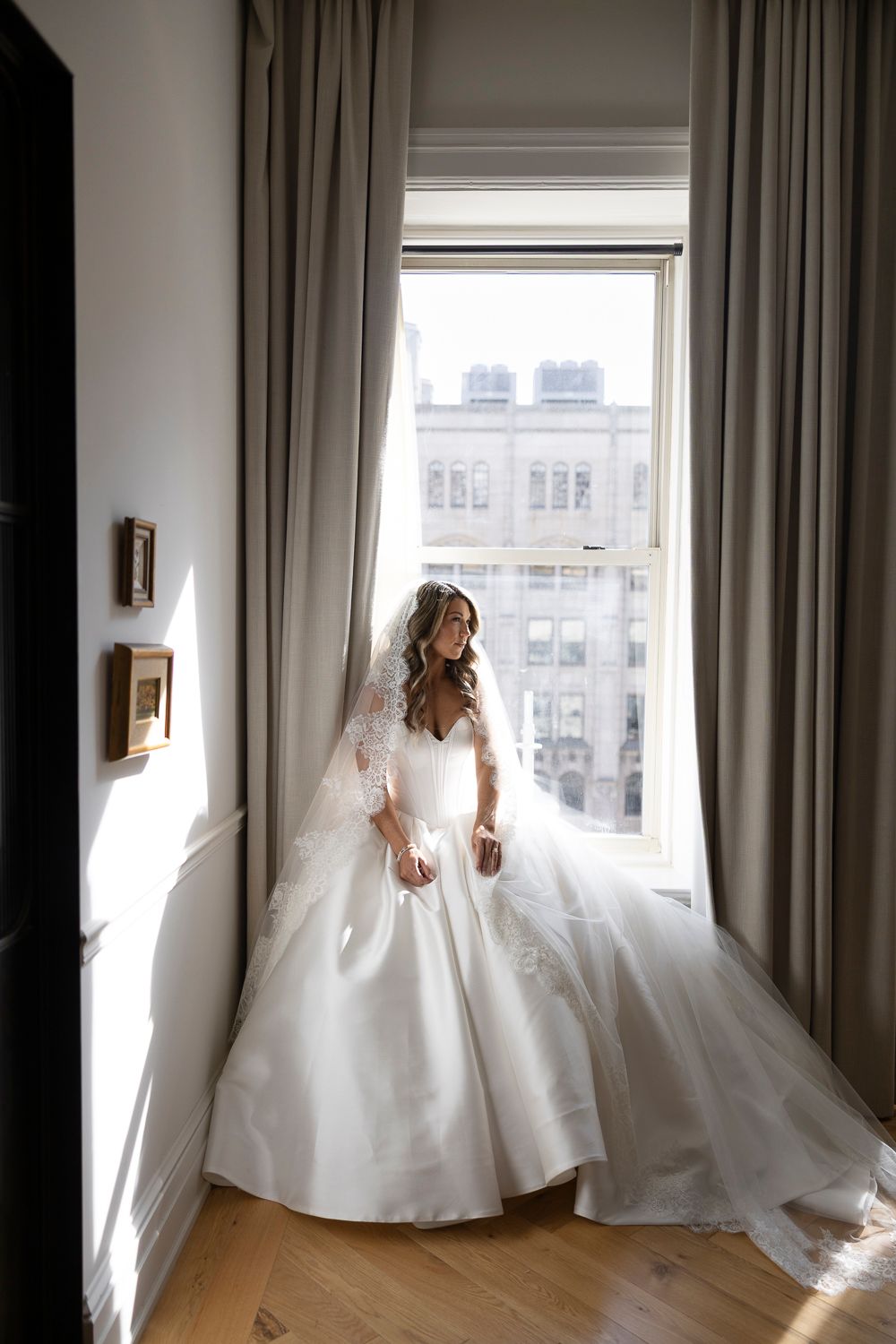 Bride in elegant white ball gown with lace sleeves stands by large window in luxurious hotel room on wedding day.