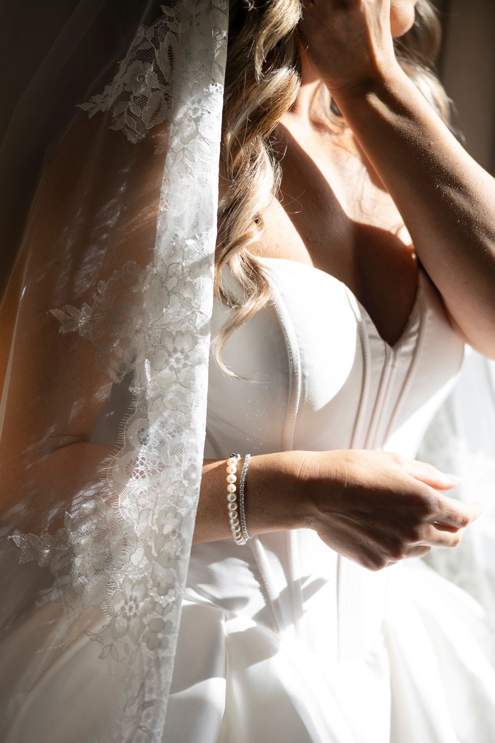 Bride in white lace wedding dress with pearl bracelet and braided hair standing by window in soft sunlight.