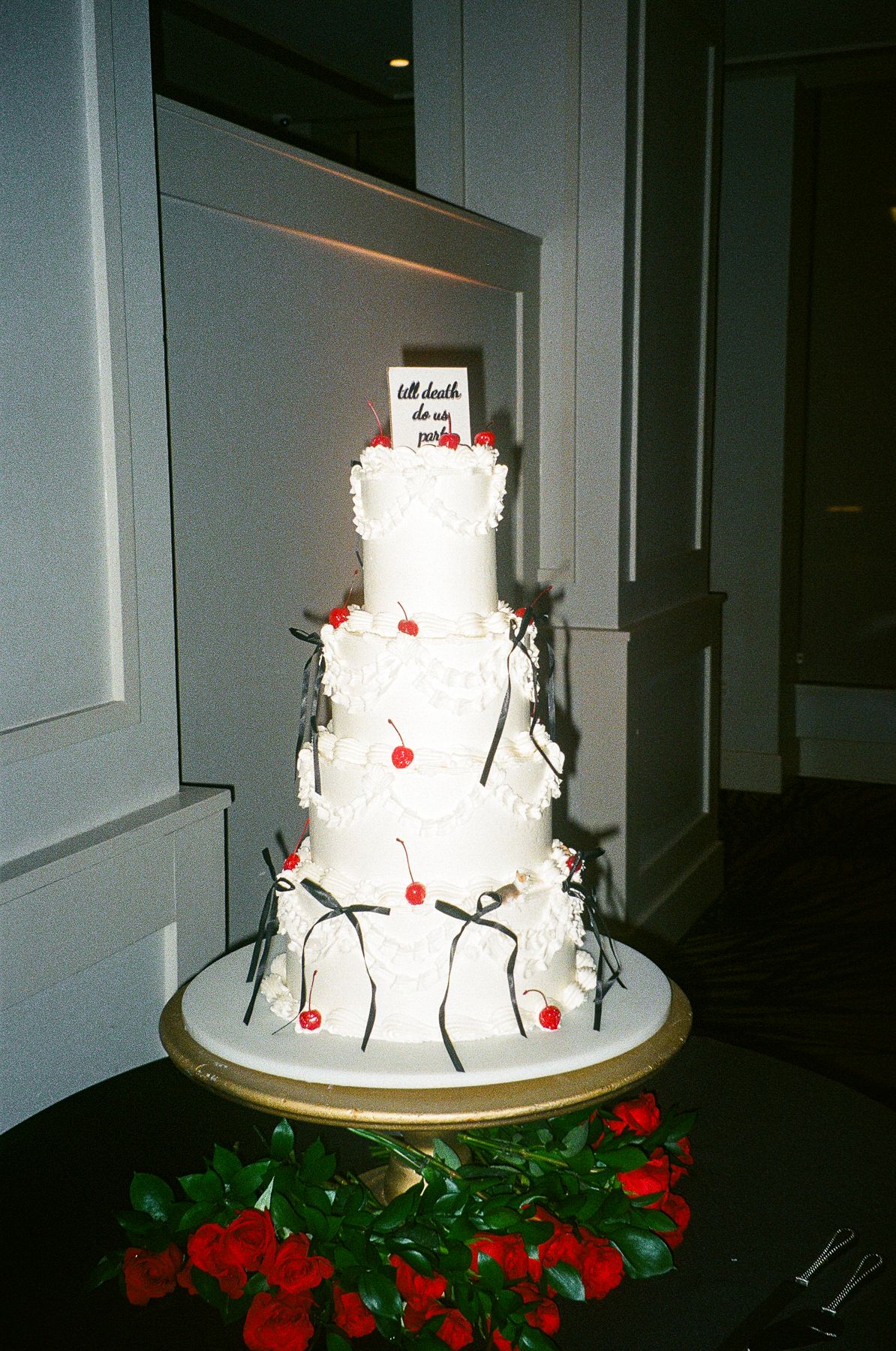 Four-tier white wedding cake with red cherries, black branches, and bows on gold stand surrounded by red roses.