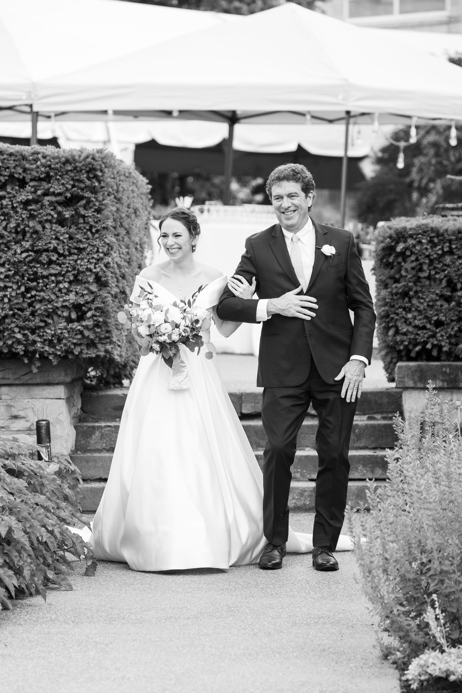 A wedding couple walking down a garden path surrounded by hedges and white umbrellas in black and white.