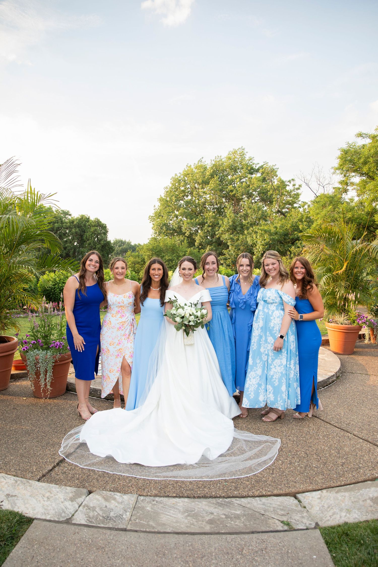 Wedding party in coordinating blue and white dresses poses together in a garden setting.