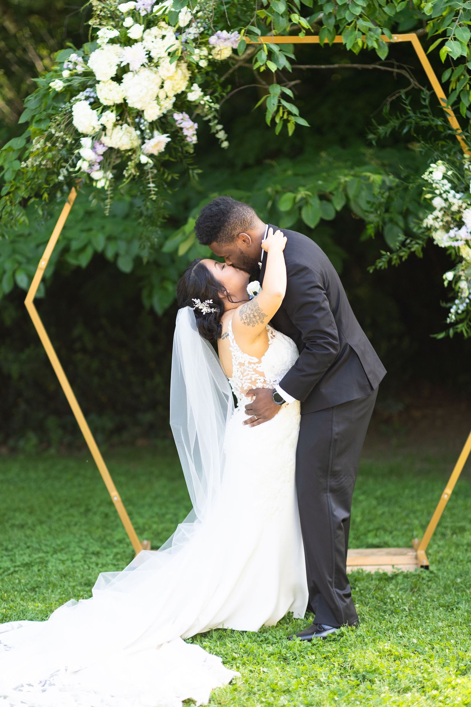 Intimate wedding ceremony moment under a floral arch on a sunny lawn.