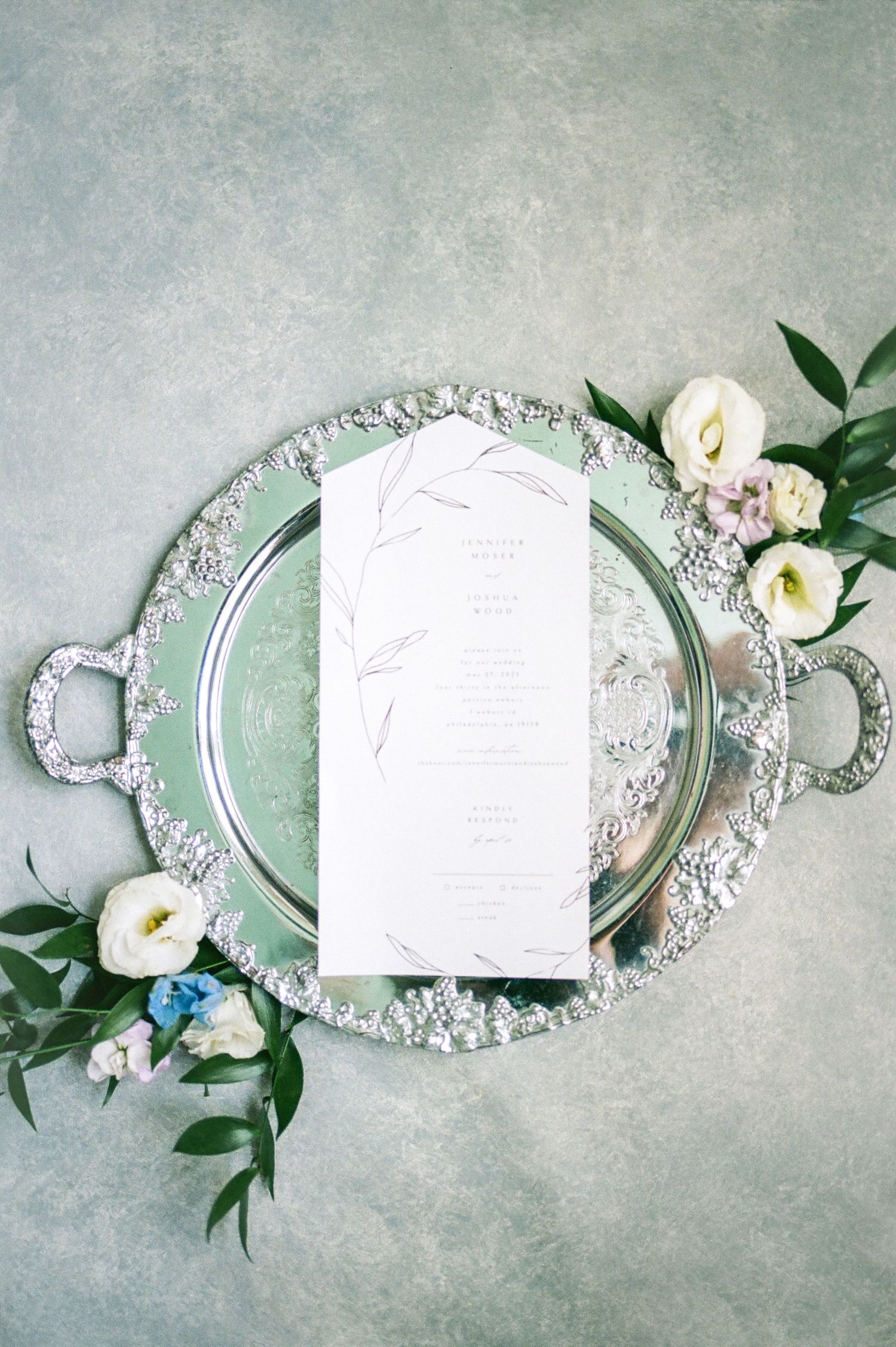 Elegant wedding invitation suite with ornate silver frames and white floral accents on sage green background.