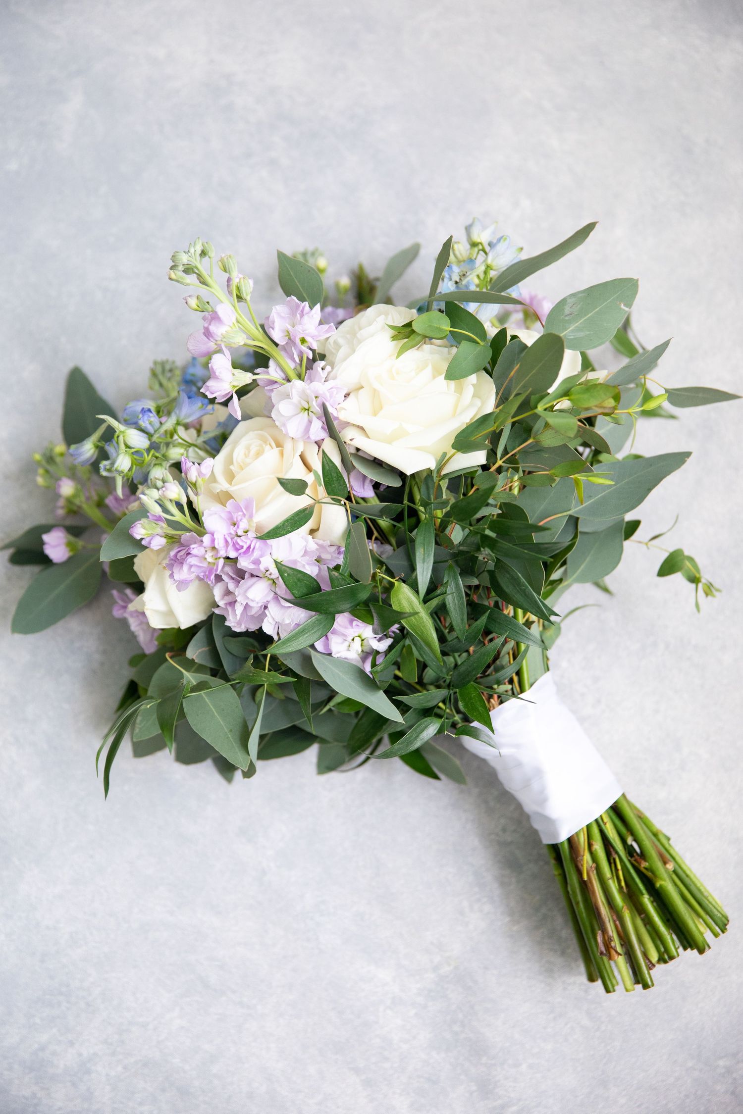 A romantic wedding bouquet with eucalyptus, white roses and purple flowers tied with white ribbon.