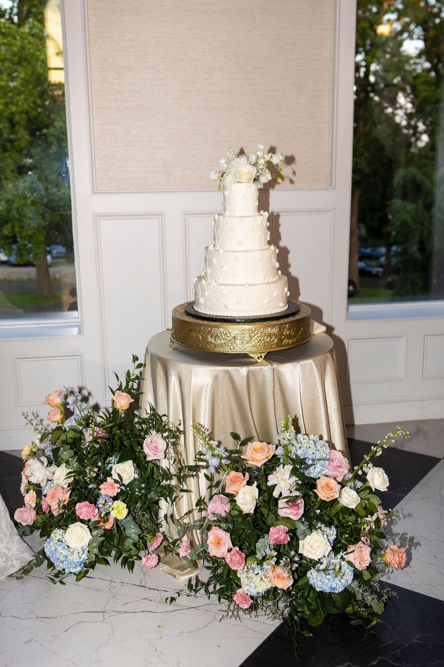 Elegant white wedding cake decorated with pink and blue flowers on a gold stand.