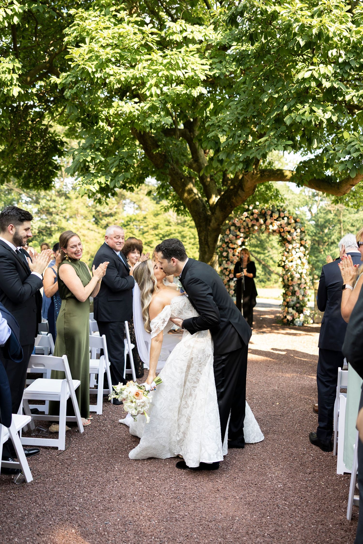 An outdoor wedding ceremony setup with white chairs under lush green trees.